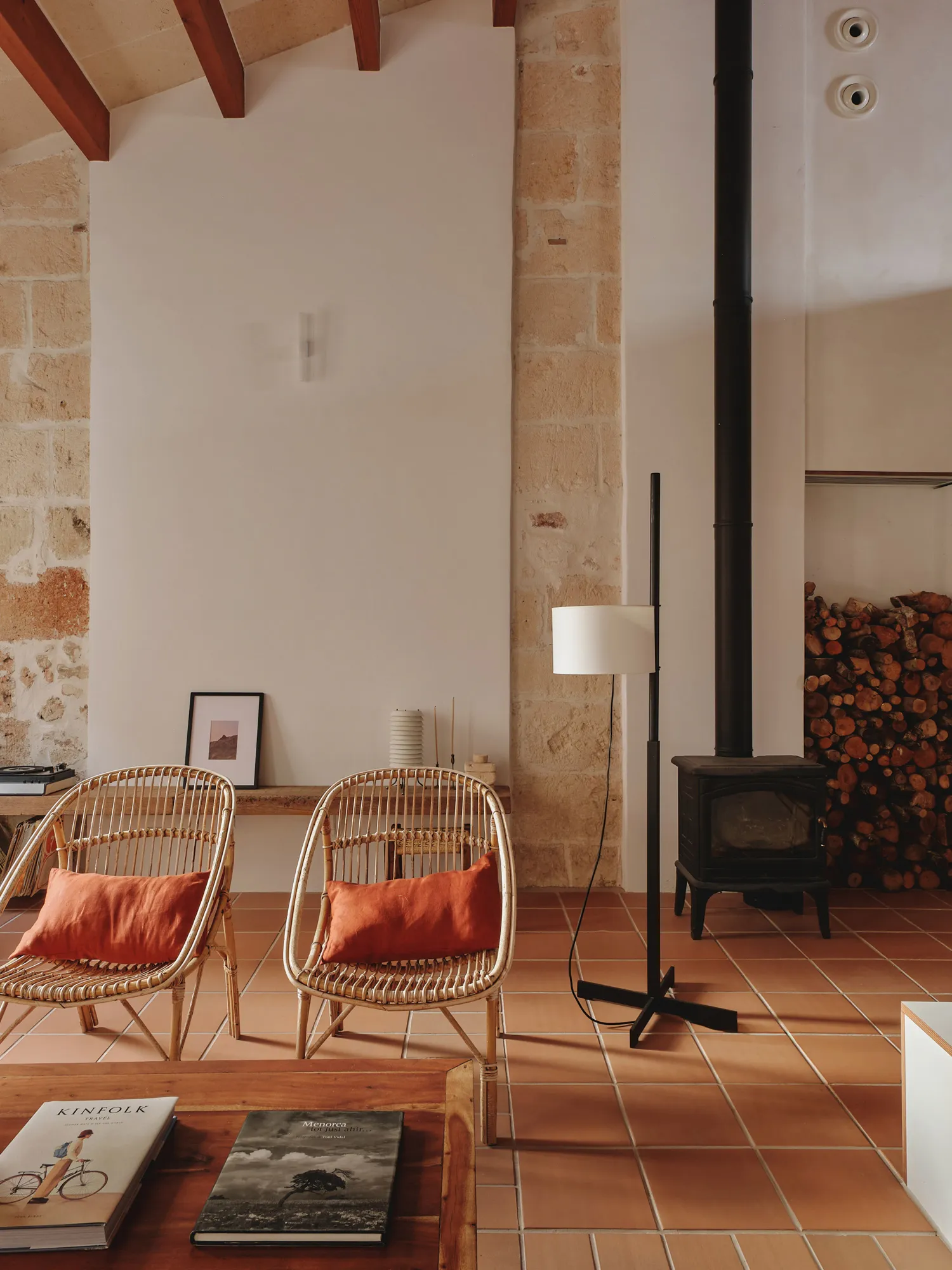 Sala de estar rústica-mediterránea con sillas de ratán, cojines naranjas, chimenea de leña y suelo terracota.
