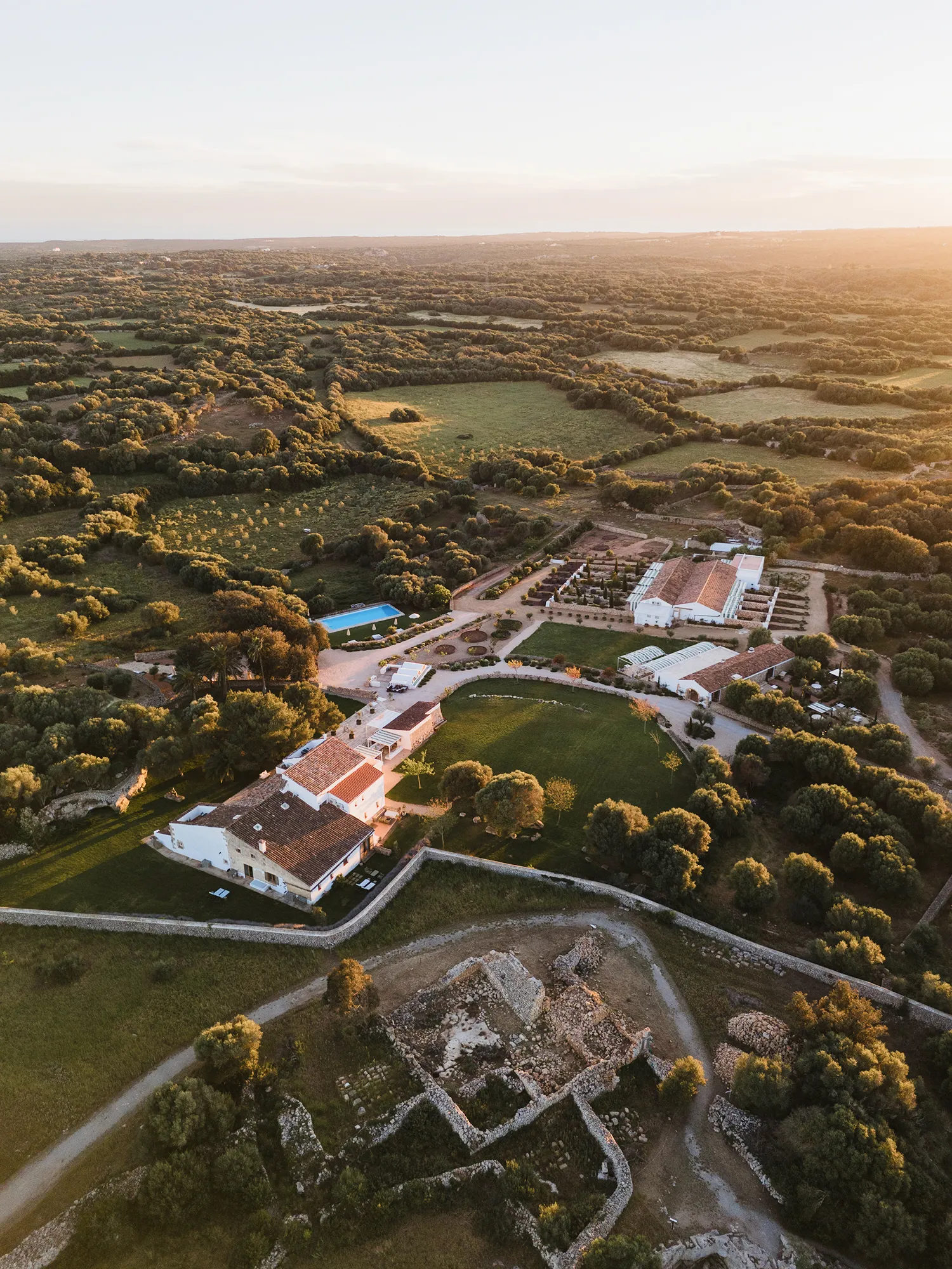 Finca rural mediterránea al atardecer, vista aérea con edificaciones, piscina y ruinas históricas en un paisaje arbolado.