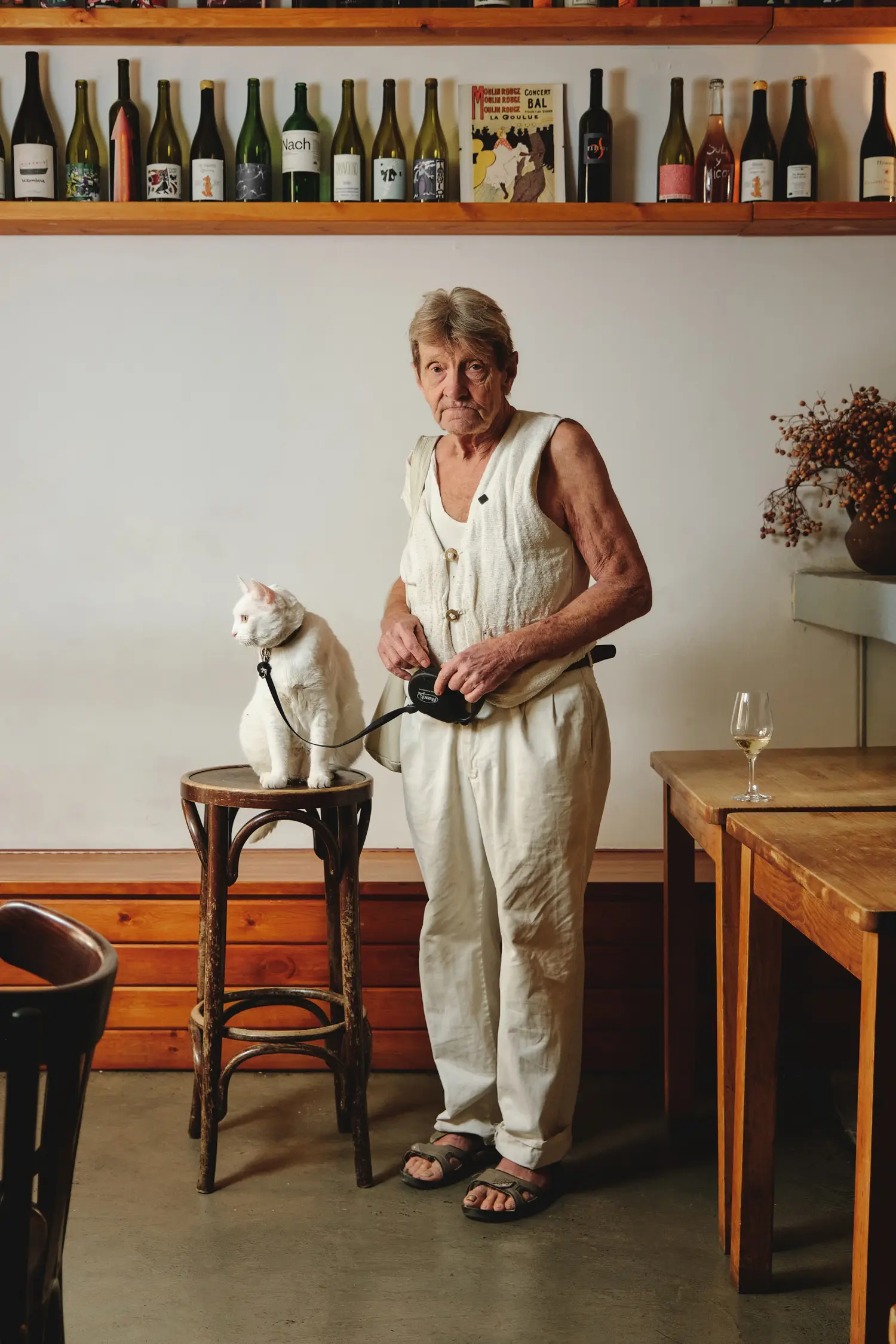 Retrato de una mujer con su gato blanco en el ambiente cálido de un bar o bodega, entre vinos y mesas de madera.