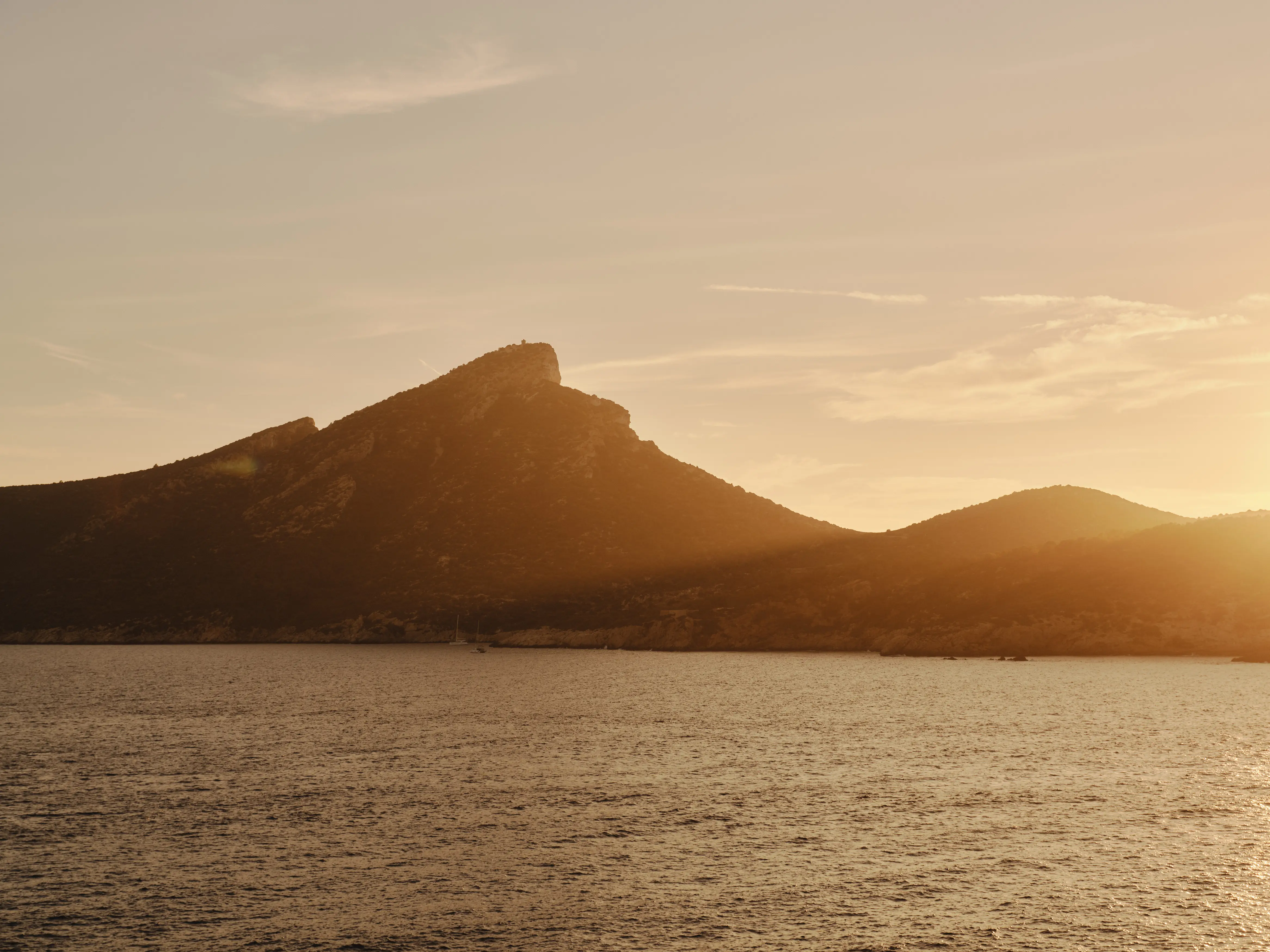 Atardecer dorado sobre montañas que se alzan desde un mar en calma, con una silueta de barco en la bahía.