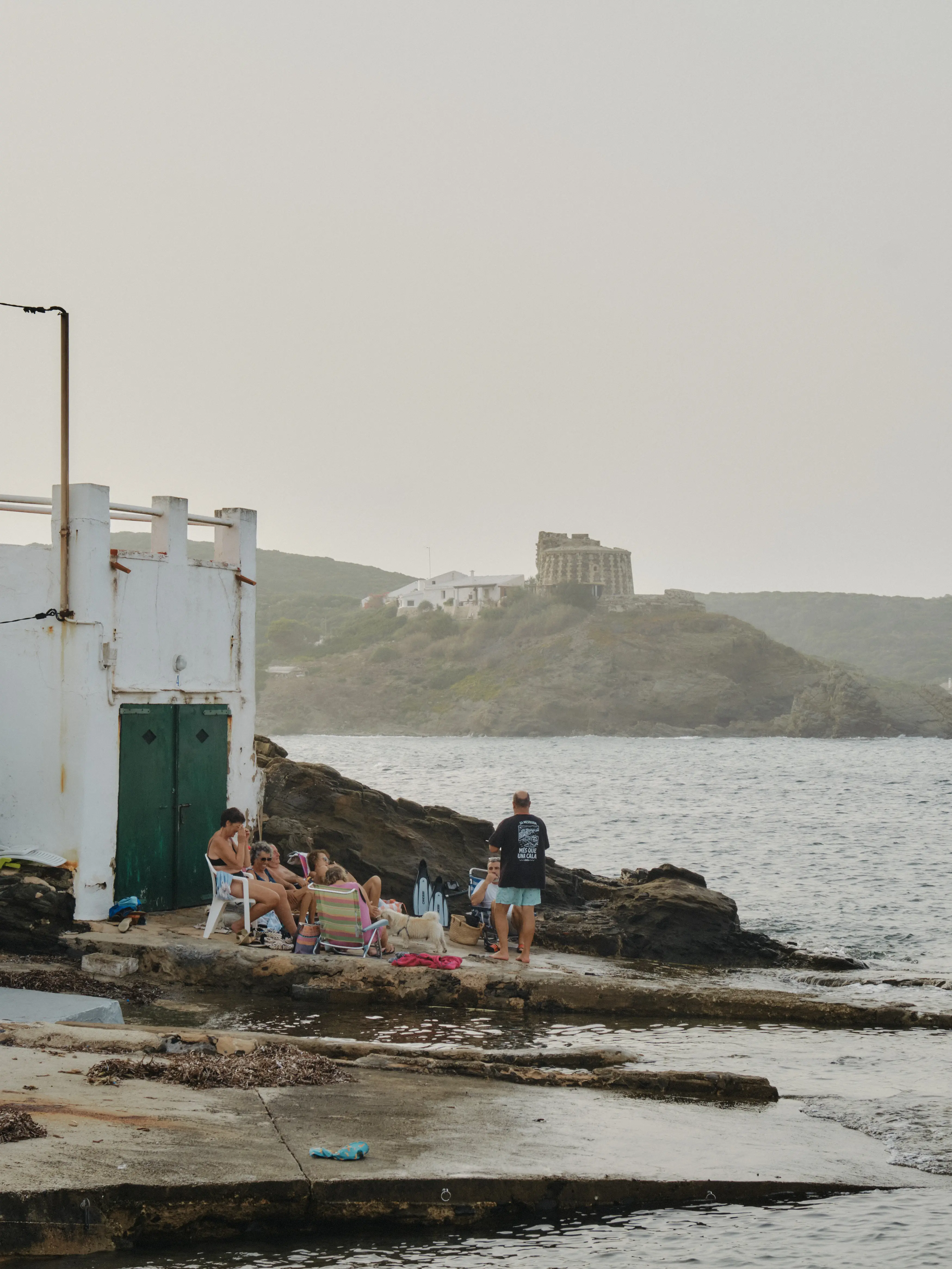 Grupo de amigos relajándose junto al mar en la costa, con un edificio histórico en la colina al fondo bajo un cielo brumoso.