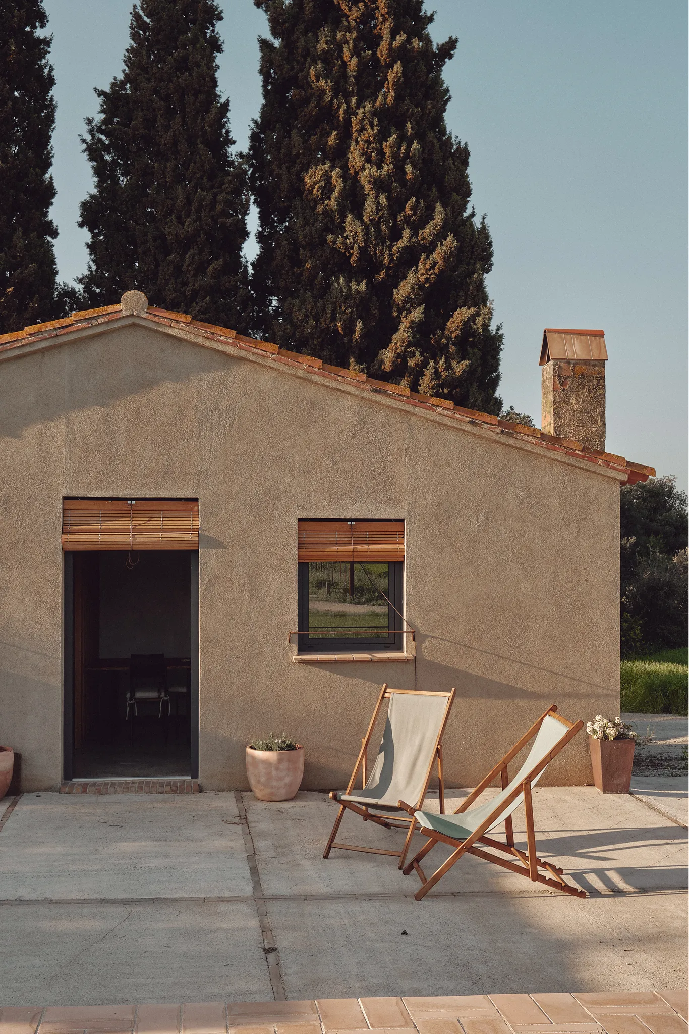 Fachada rústica mediterránea con patio soleado, dos sillas de lona y cipreses altos.