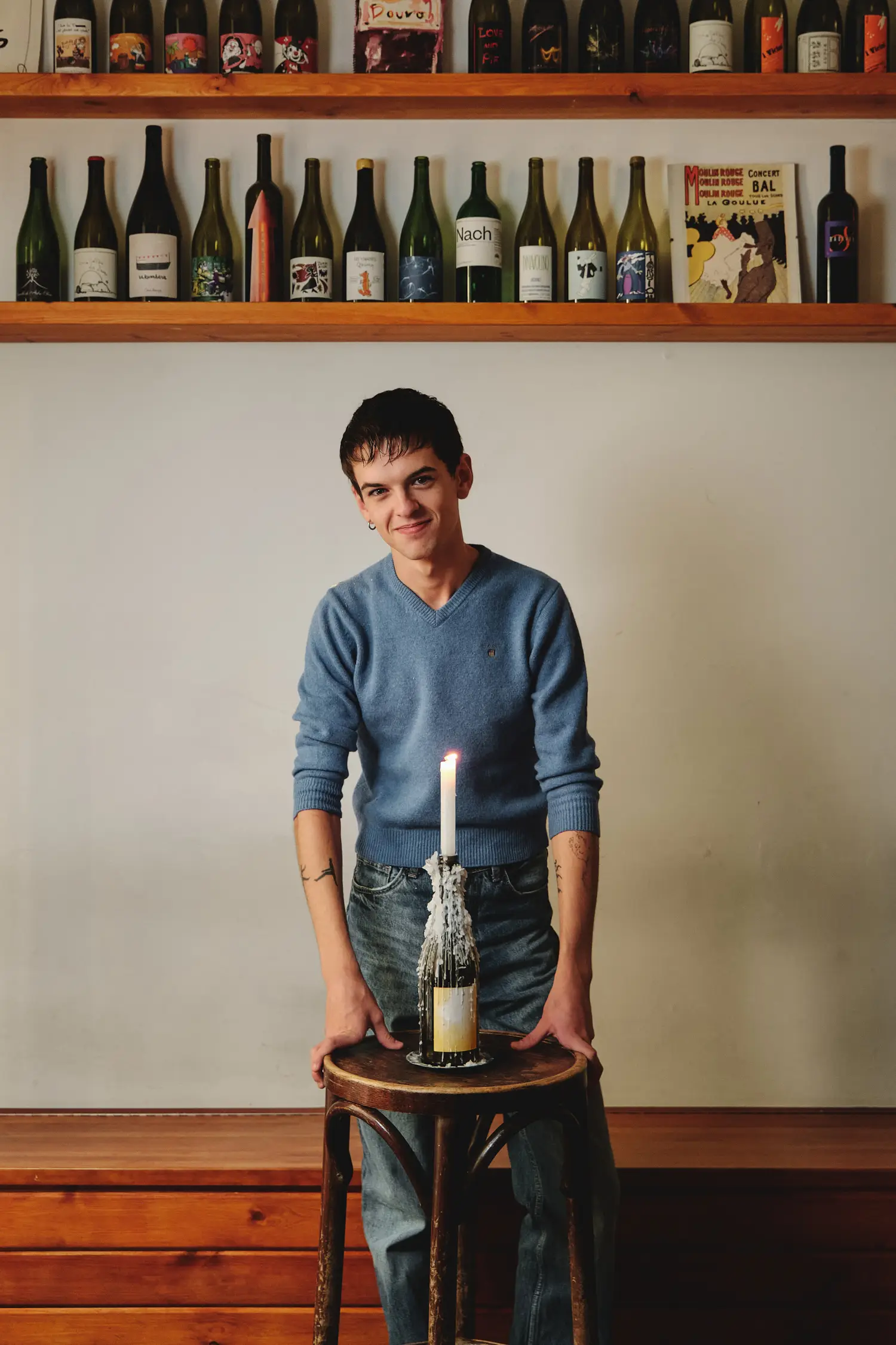 Joven sonriente en suéter azul sujeta taburete con botella de vino y vela, rodeado de estantes de vino.