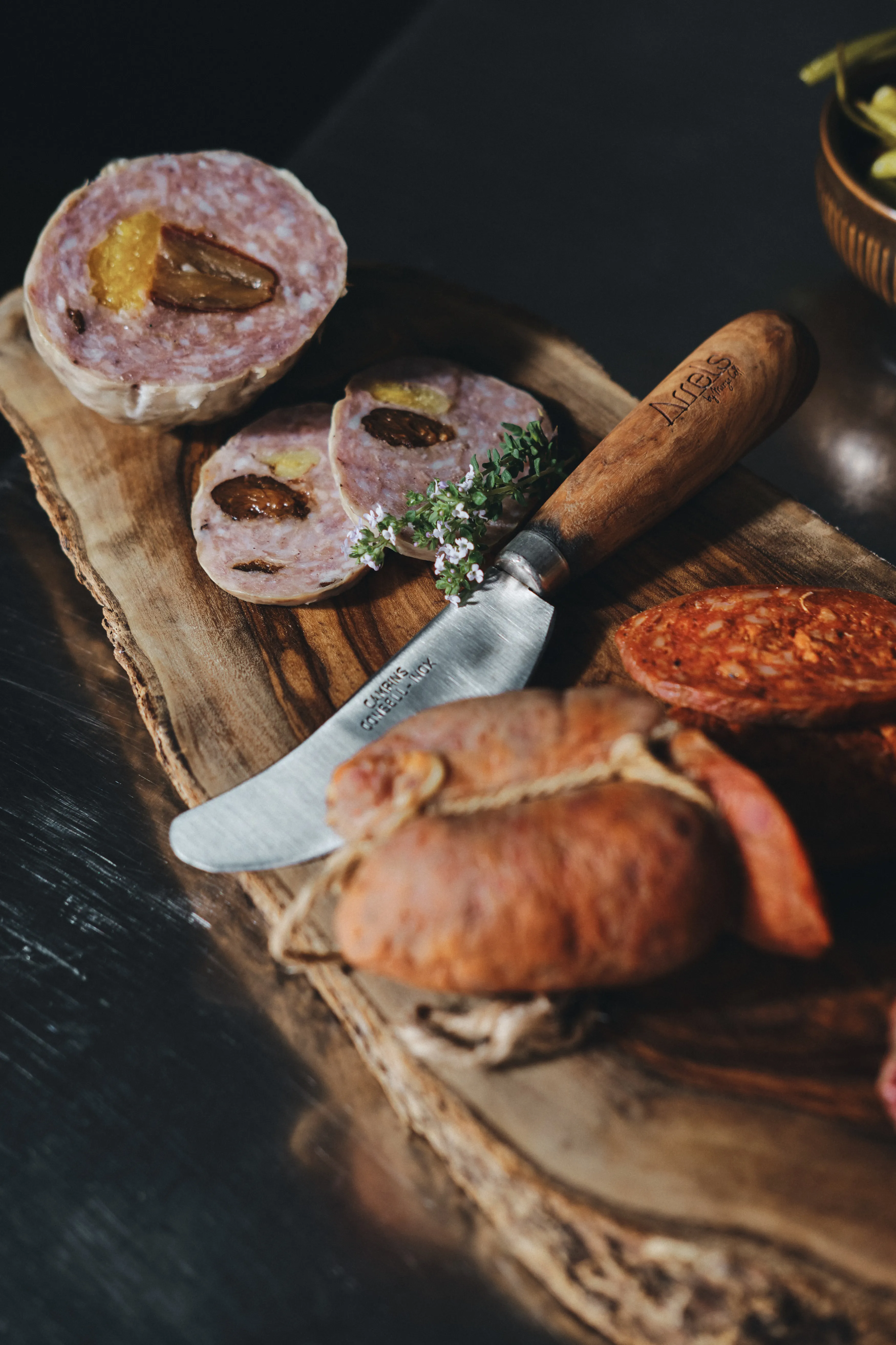 Selección gourmet de embutidos en tabla de madera, destacando salami con frutos secos y chorizo, con cuchillo y hierbas.