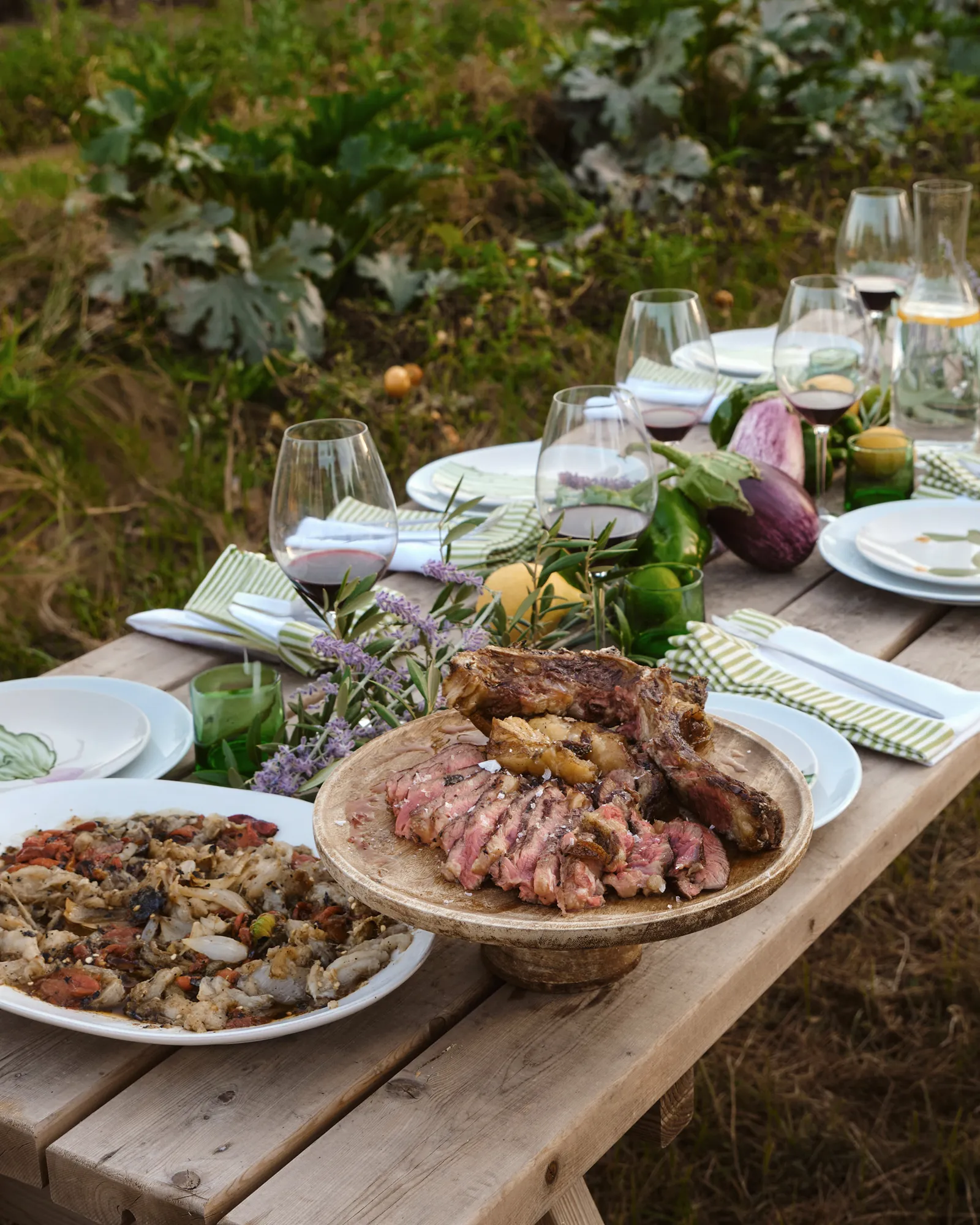 Cena campestre mediterránea sobre mesa de madera rústica, con chuletón, marisco, copas de vino y decoración natural.