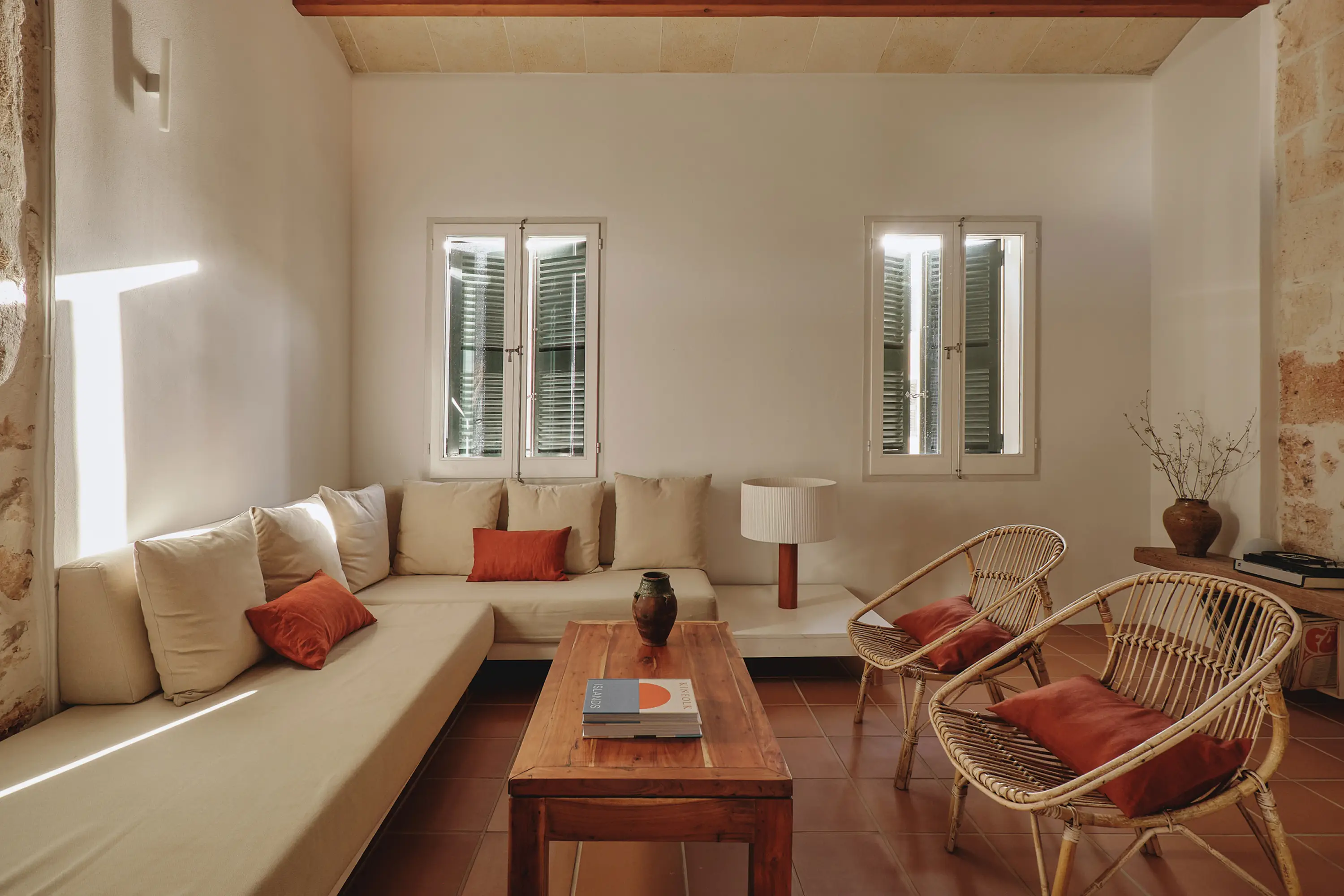 Salón luminoso de estilo mediterráneo con sofá modular, sillones de ratán, mesa de madera y cojines terracota.