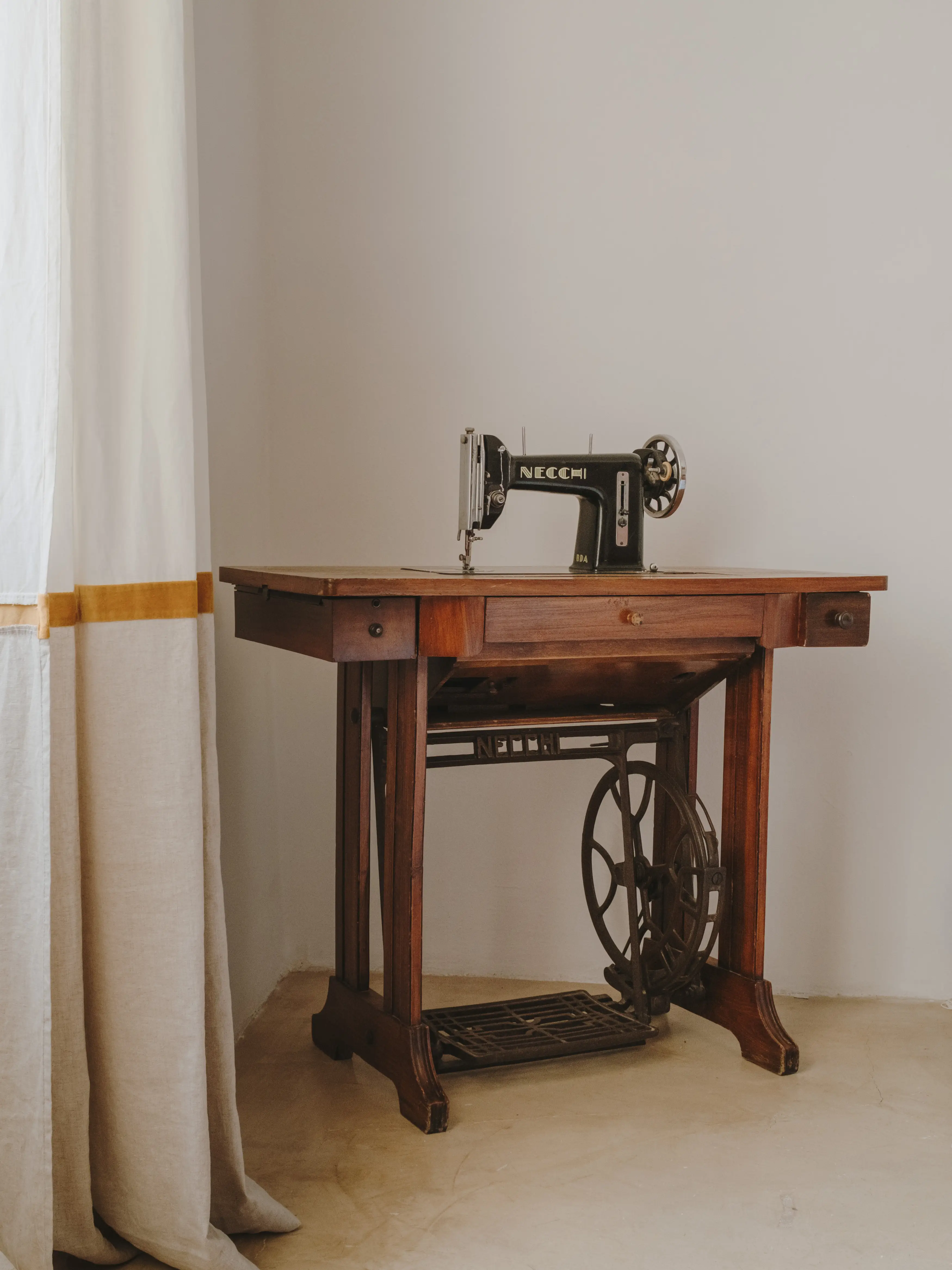 Antigua máquina de coser Necchi sobre mesa de madera con pie de hierro, junto a cortina clara en un interior luminoso.
