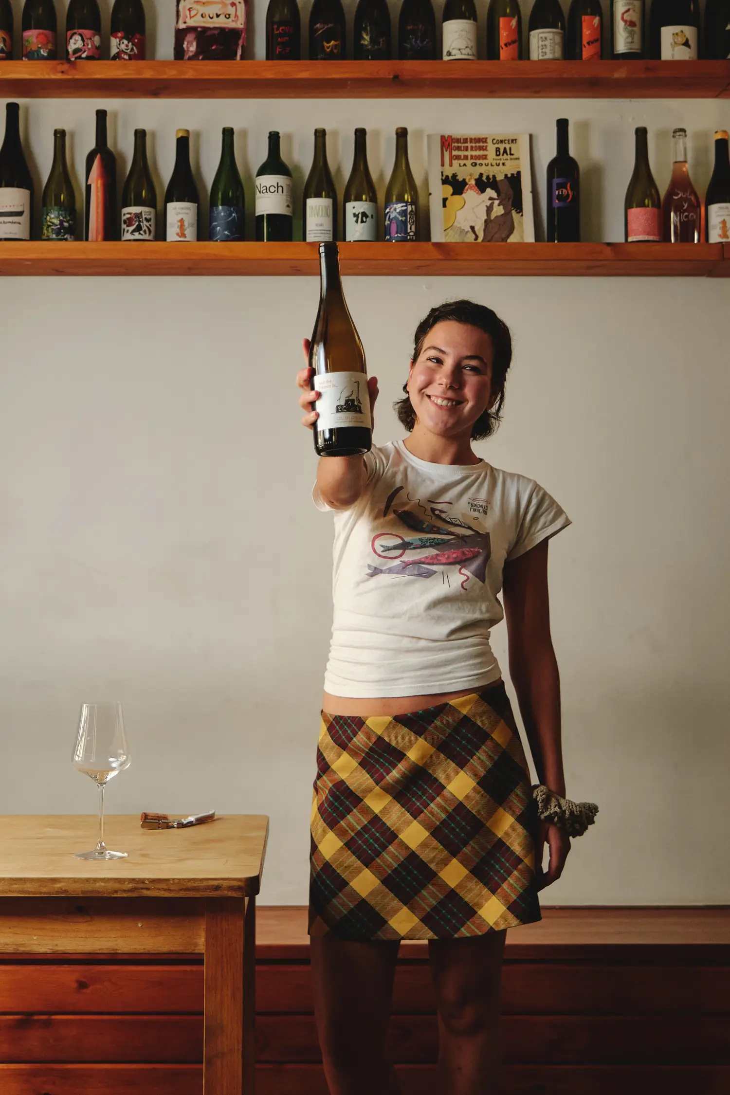 Joven sonriente mostrando una botella de vino, rodeada de estanterías llenas, en un acogedor espacio.