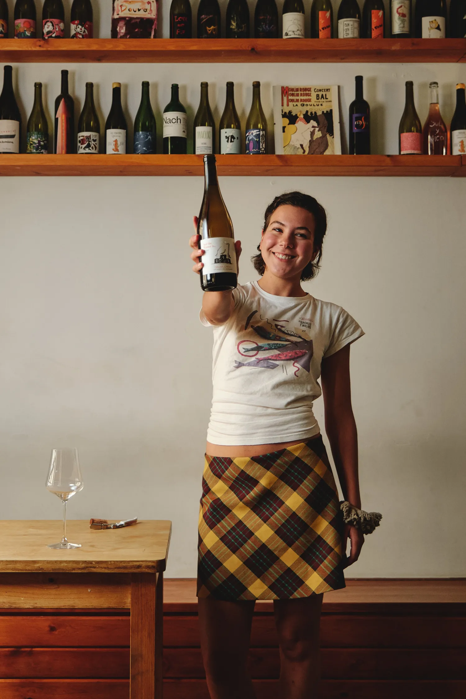 Joven sonriente mostrando una botella de vino, rodeada de estanterías llenas, en un acogedor espacio.
