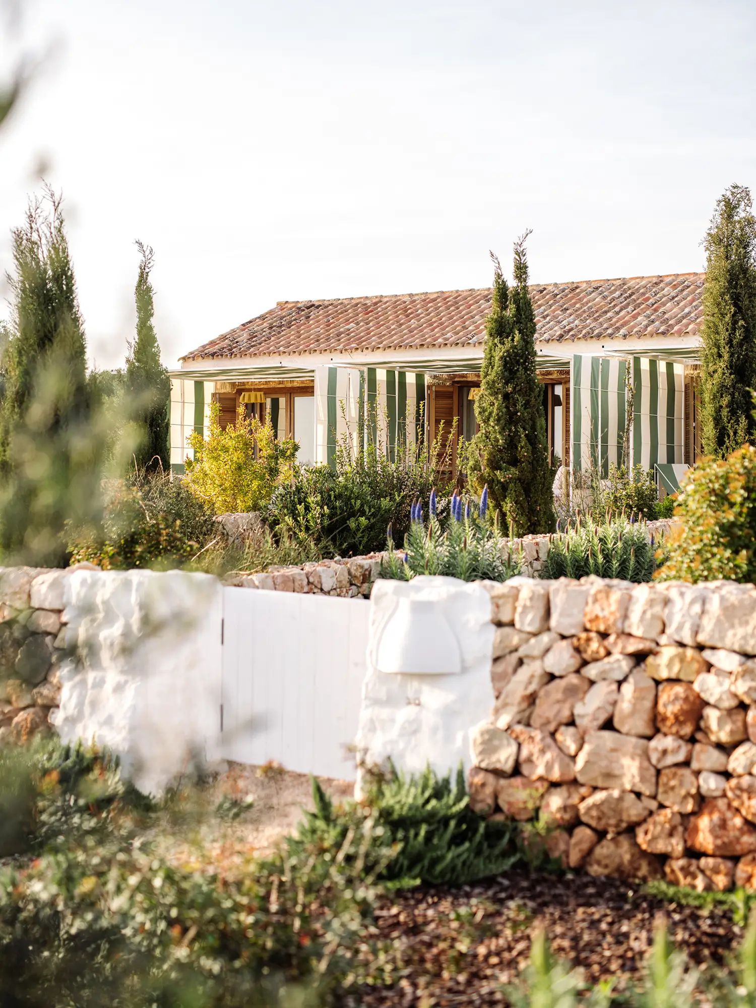 Idílica casa mediterránea de tejado rojo y toldos verdes en un jardín soleado con muro de piedra y vegetación.