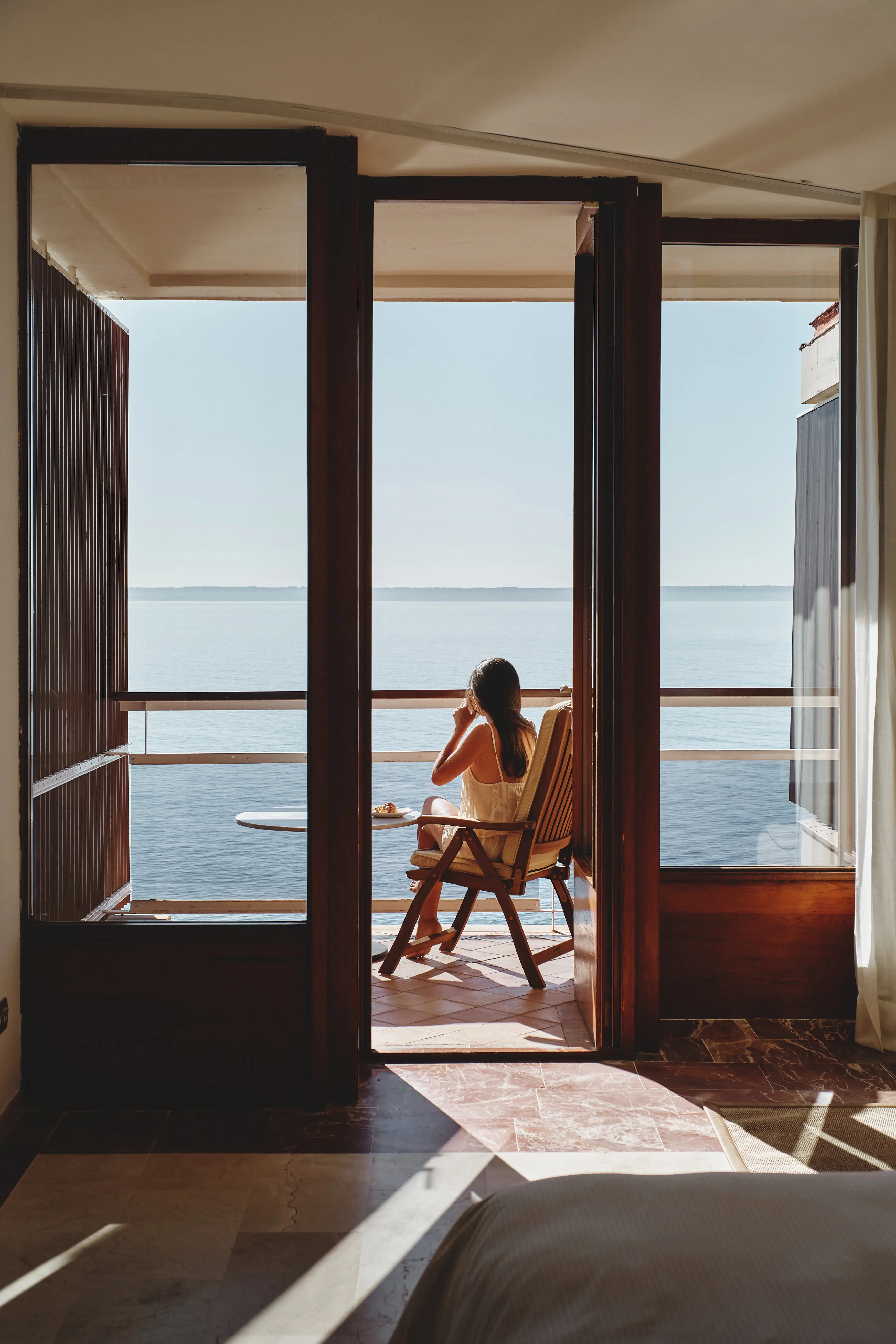 Mujer en un balcón soleado con vistas al mar infinito, disfrutando de un desayuno relajado.