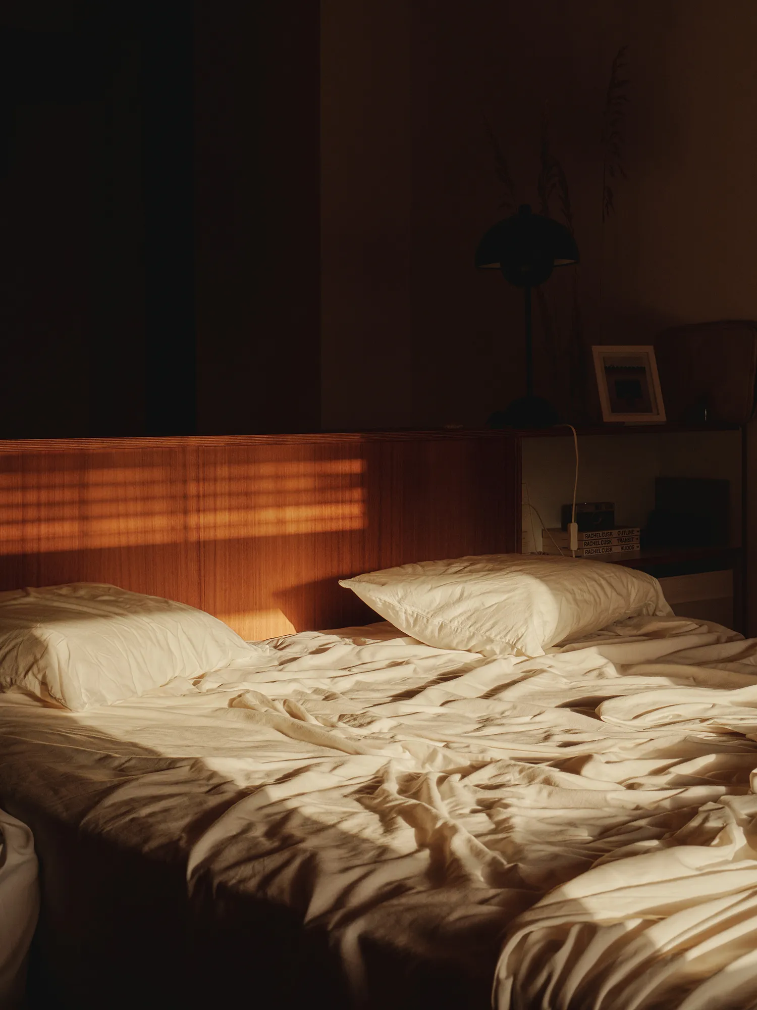 Cama con sábanas blancas y almohadas, iluminada por rayas de sol que entran por la ventana en una habitación serena.