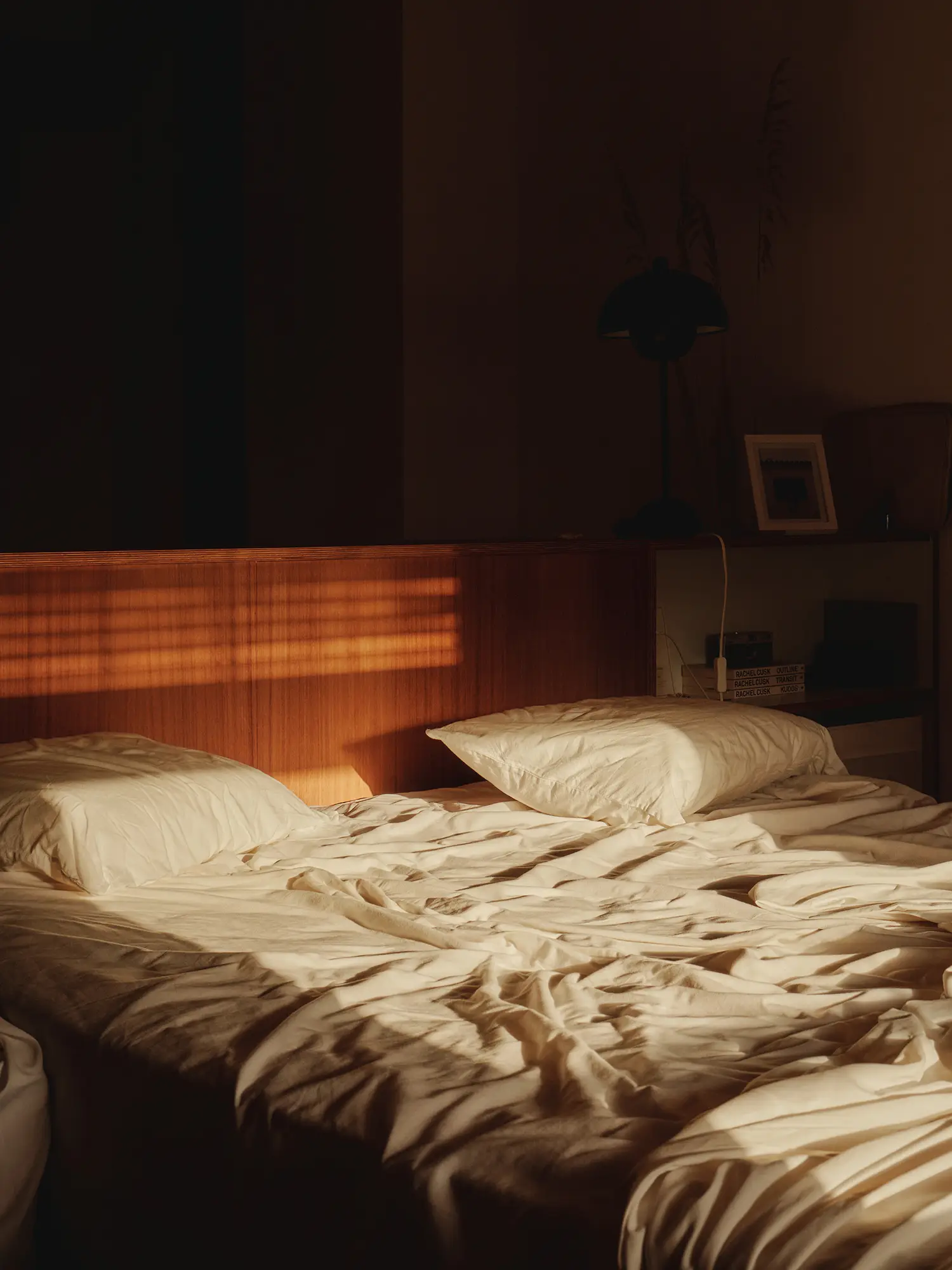 Cama con sábanas blancas y almohadas, iluminada por rayas de sol que entran por la ventana en una habitación serena.