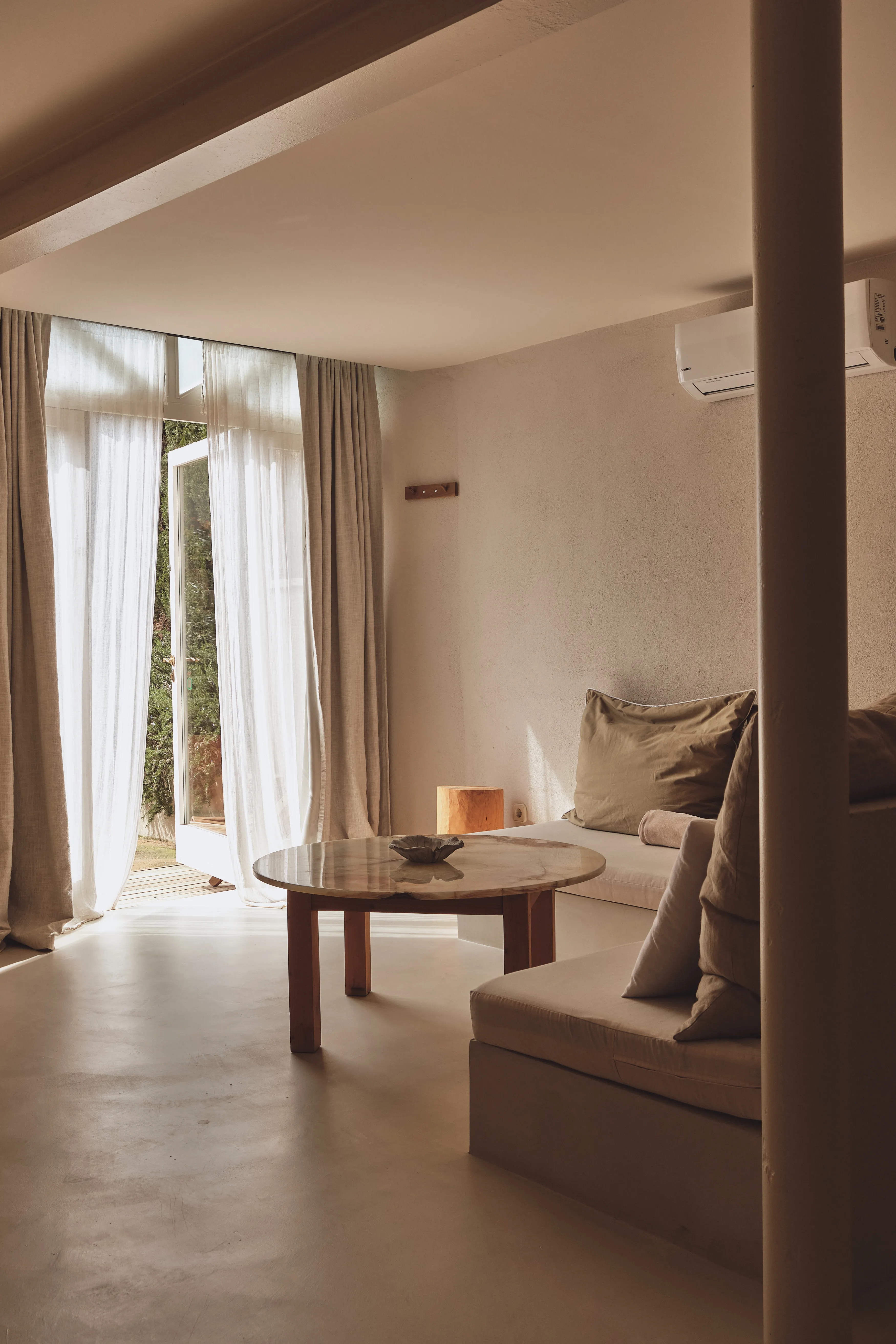 Salón interior minimalista con sofá de obra, mesa redonda de mármol y grandes ventanales que dan a un jardín verde.