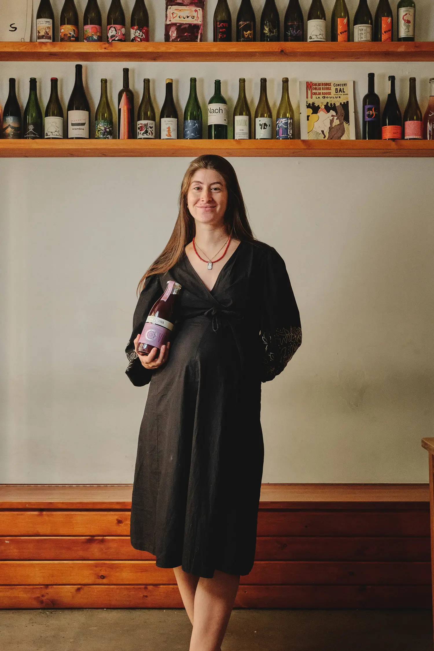 Mujer embarazada sonriente sosteniendo una botella de vino en un ambiente cálido con estantes llenos de botellas.