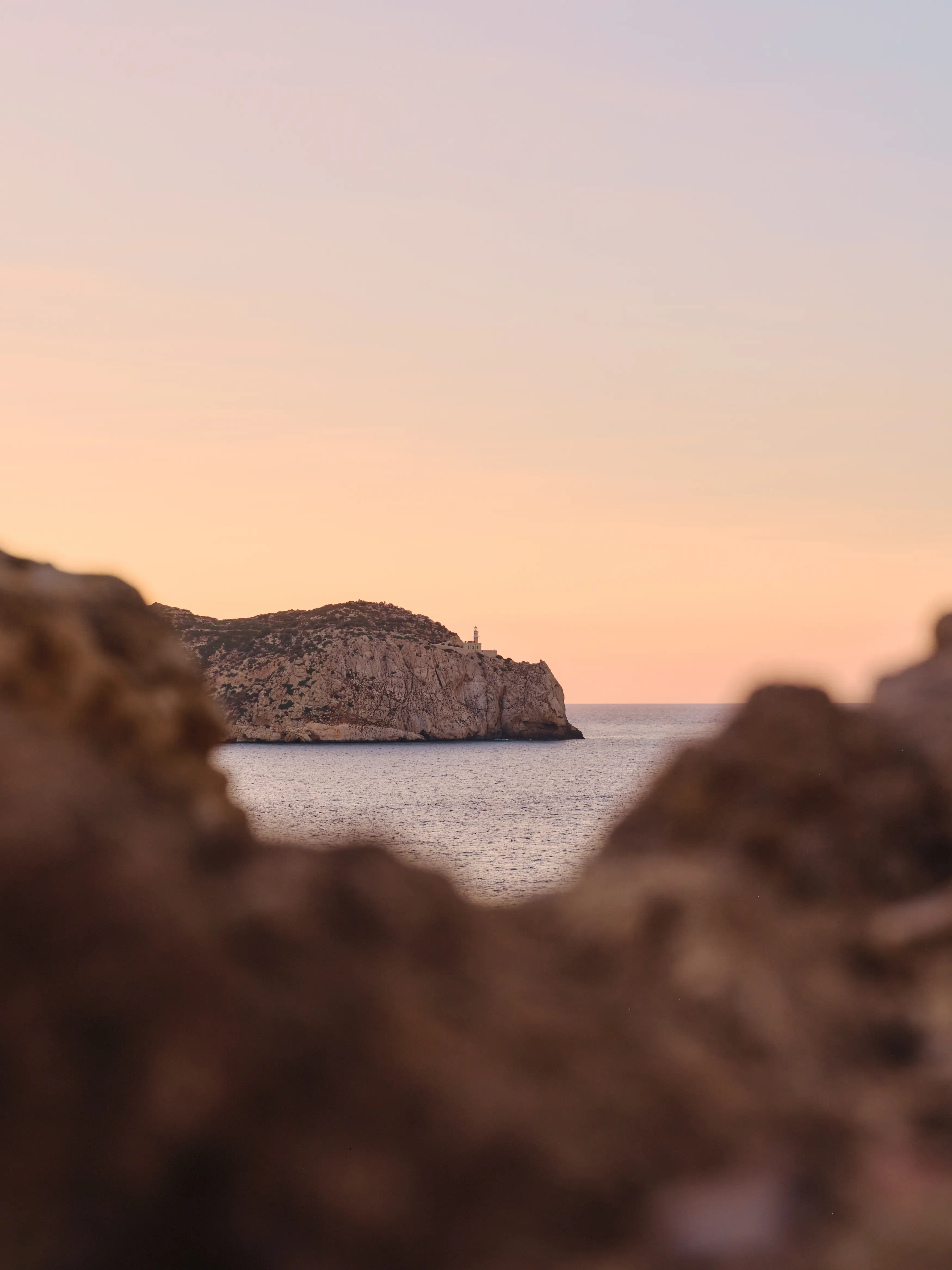 Silueta de un promontorio rocoso con un faro al atardecer, enmarcado por rocas en primer plano y mar en calma.