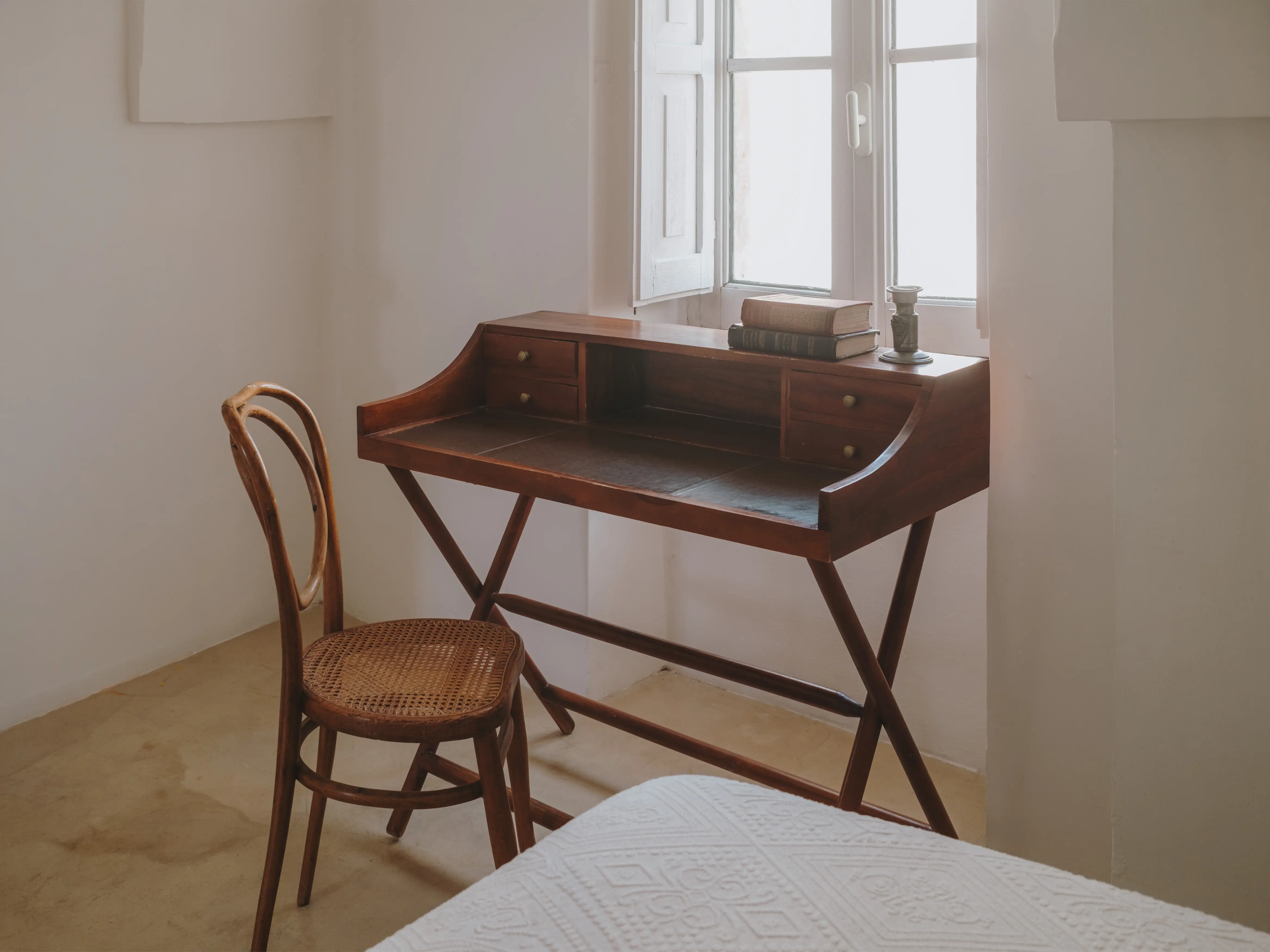 Rincón de dormitorio sereno con escritorio de madera antigua junto a ventana, silla de ratán y cama con colcha blanca.