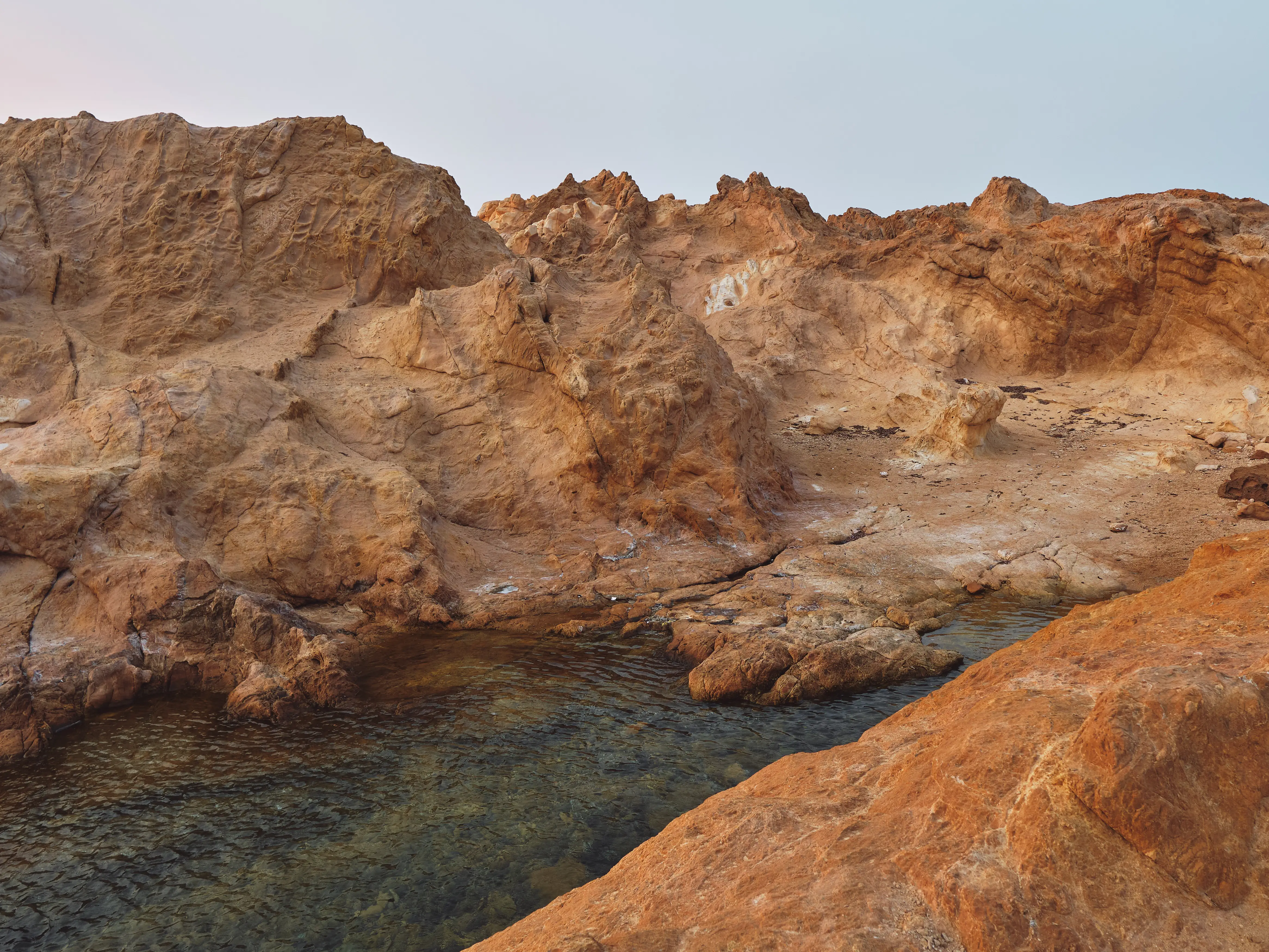 Formaciones rocosas de color rojizo junto a aguas cristalinas en una cala mediterránea bajo un cielo despejado.