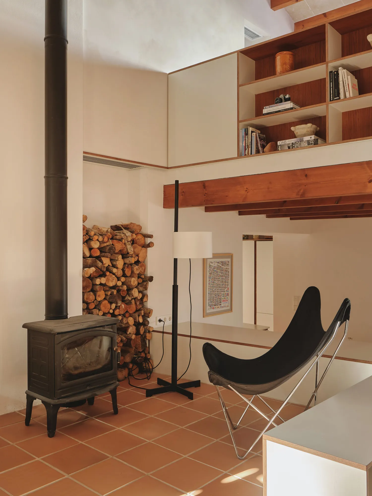 Acogedor salón con estufa de leña, pila de troncos y mobiliario de diseño sobre suelo de terracota en una casa mediterránea.