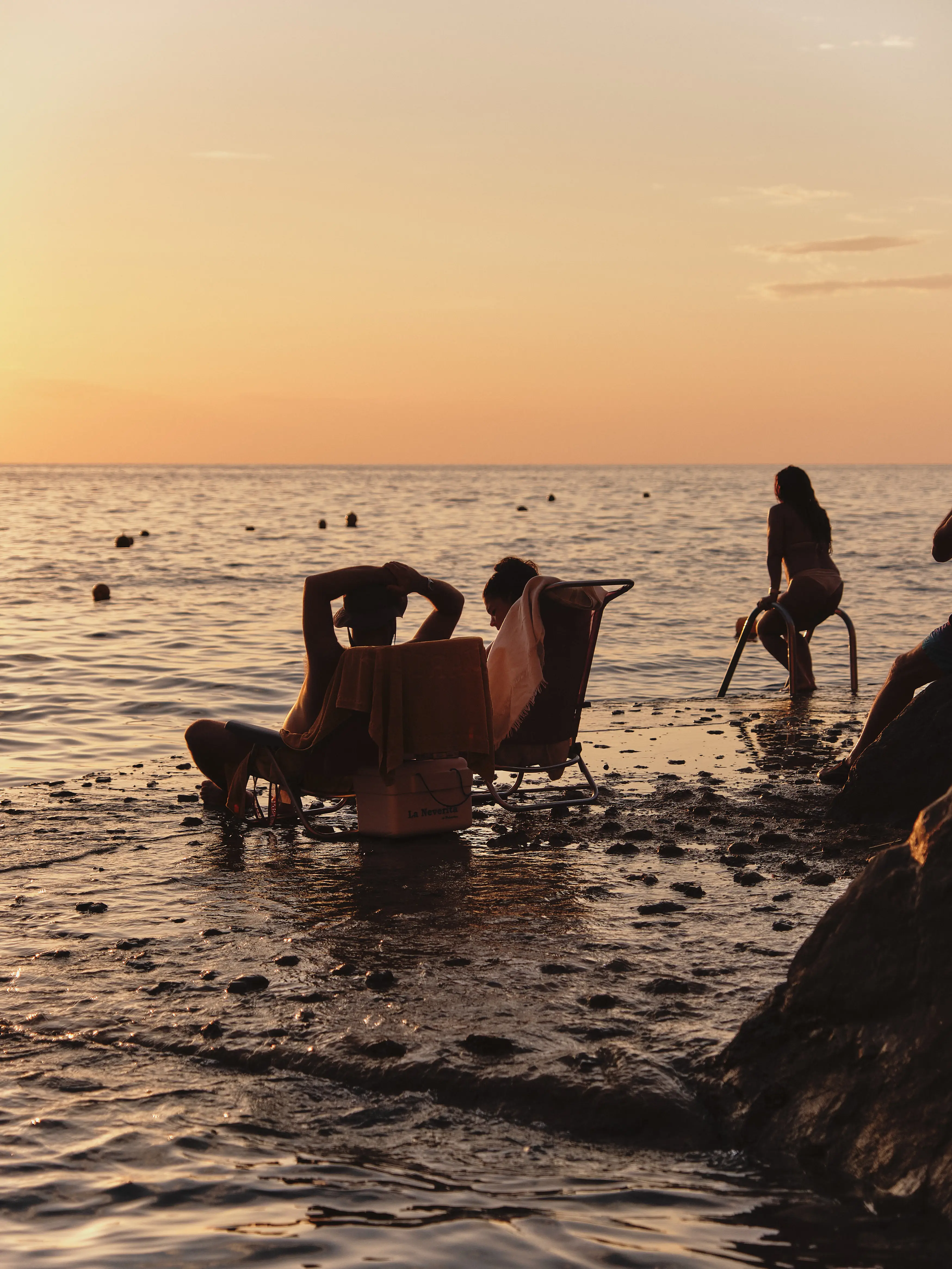 Siluetas de bañistas relajándose en la orilla del mar al atardecer, disfrutando del ambiente tranquilo y la luz dorada.