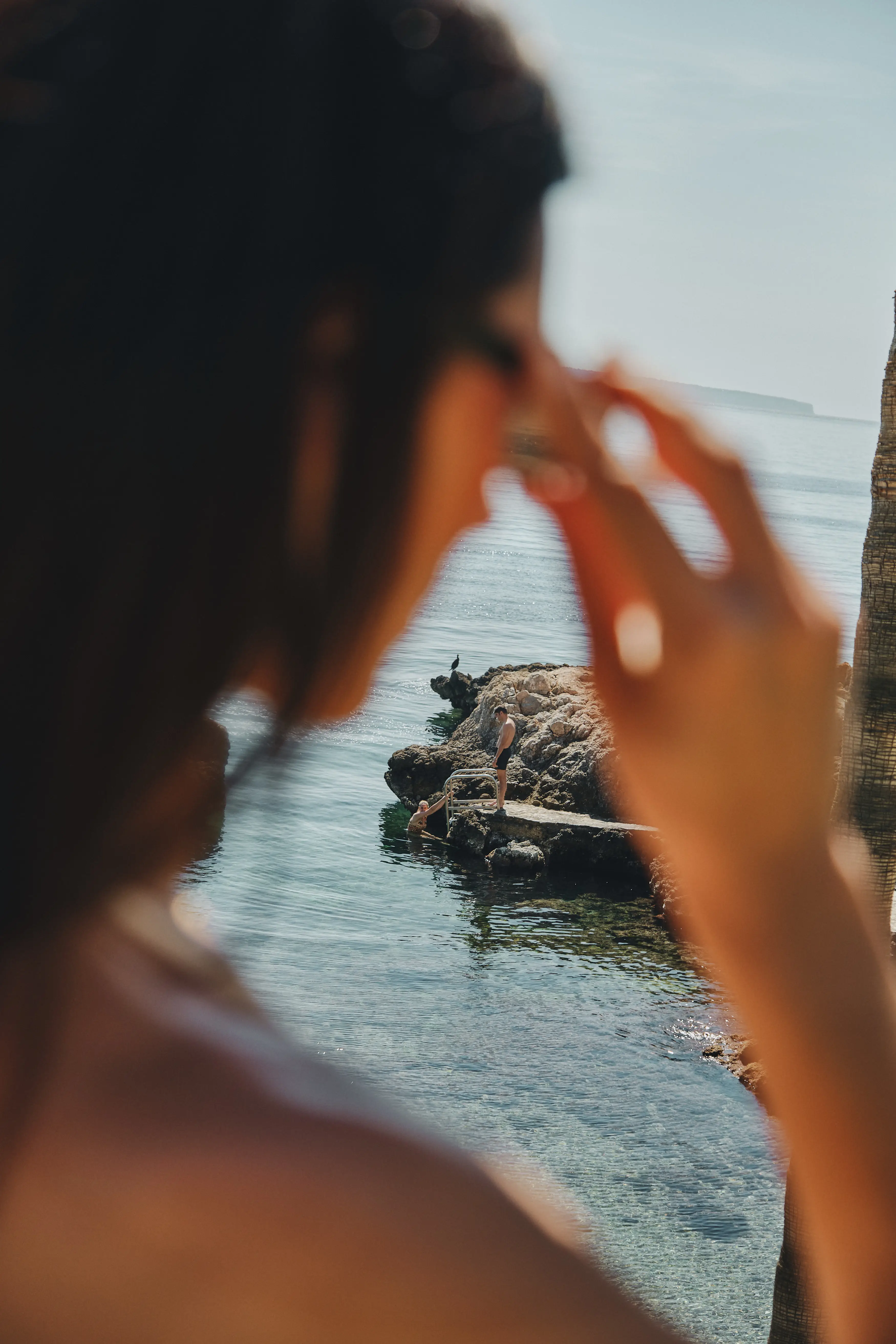 Persona observando a dos bañistas en una cala rocosa mediterránea bajo el sol.