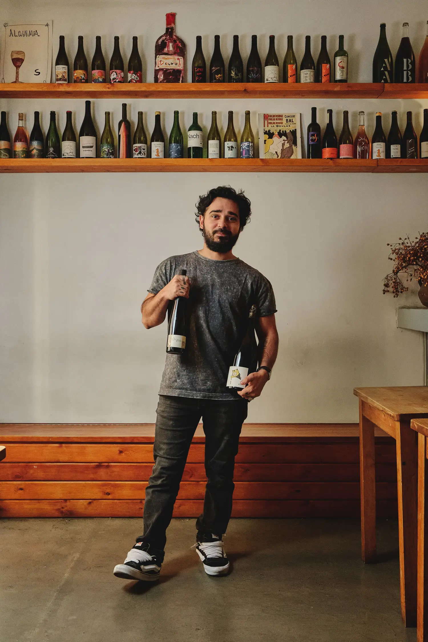 Sommelier sonriente muestra dos botellas de vino en un ambiente rústico con estantes llenos de vinos.