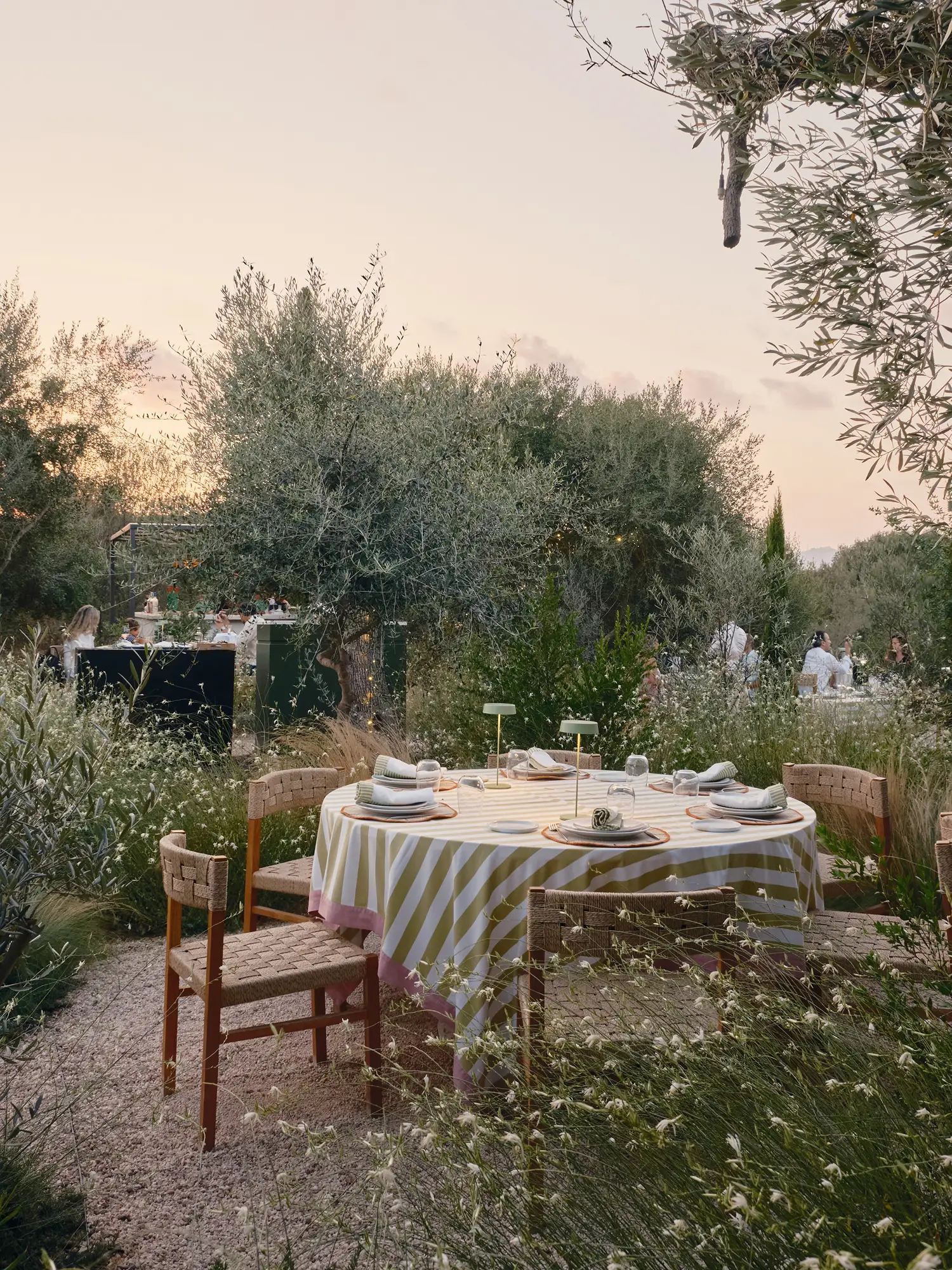 Elegante mesa de comedor exterior en jardín mediterráneo al atardecer, con mantel rayado y sillas de fibras naturales.