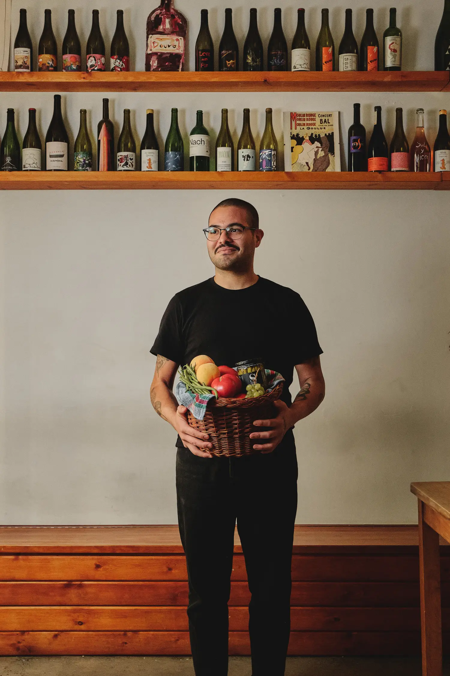 Hombre sonriente con gafas y camiseta negra sosteniendo una cesta de frutas y verduras frente a una estantería de vinos.