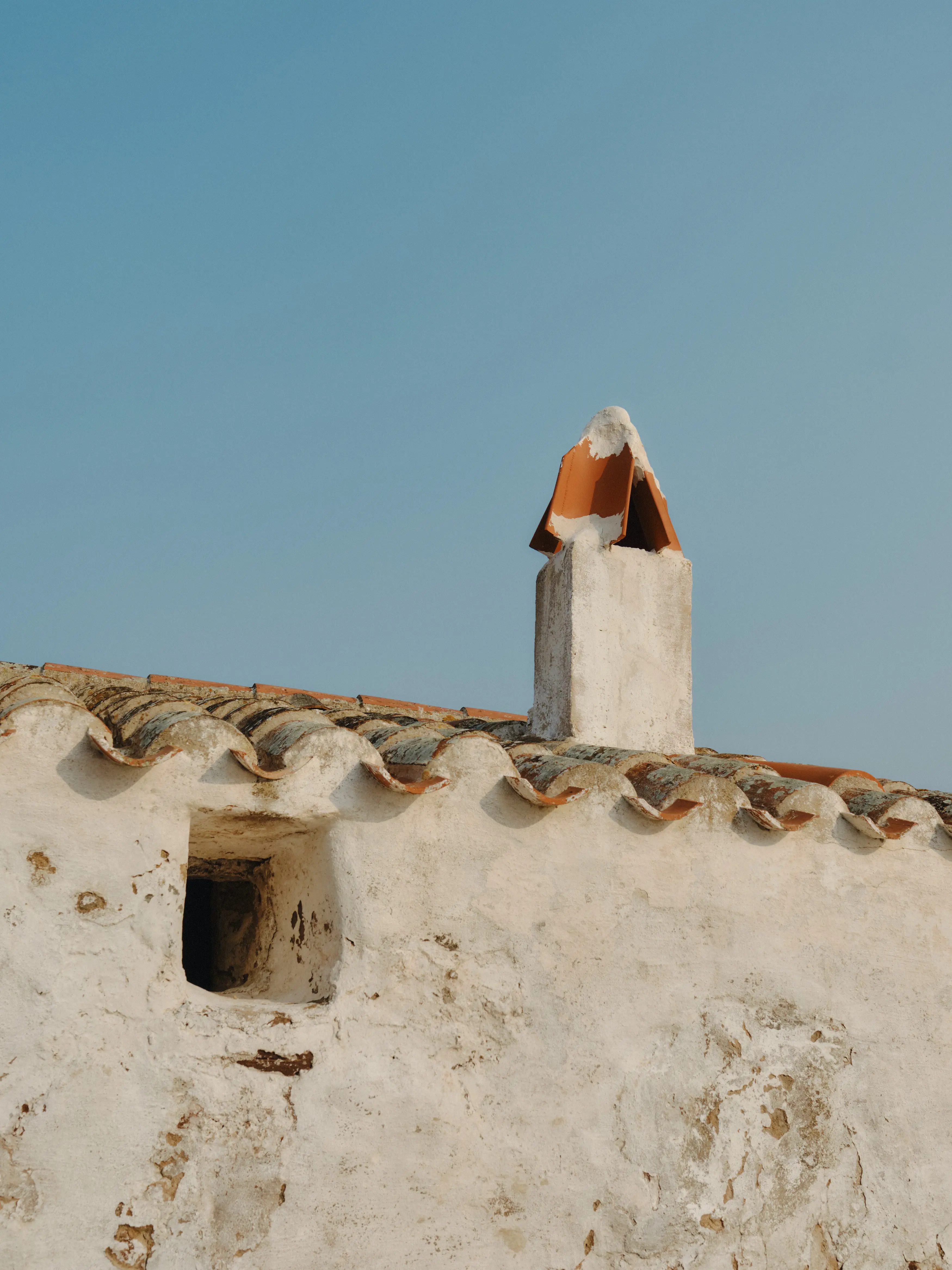 Detalle de una fachada rústica encalada con tejas de terracota y una chimenea contra un cielo azul claro.