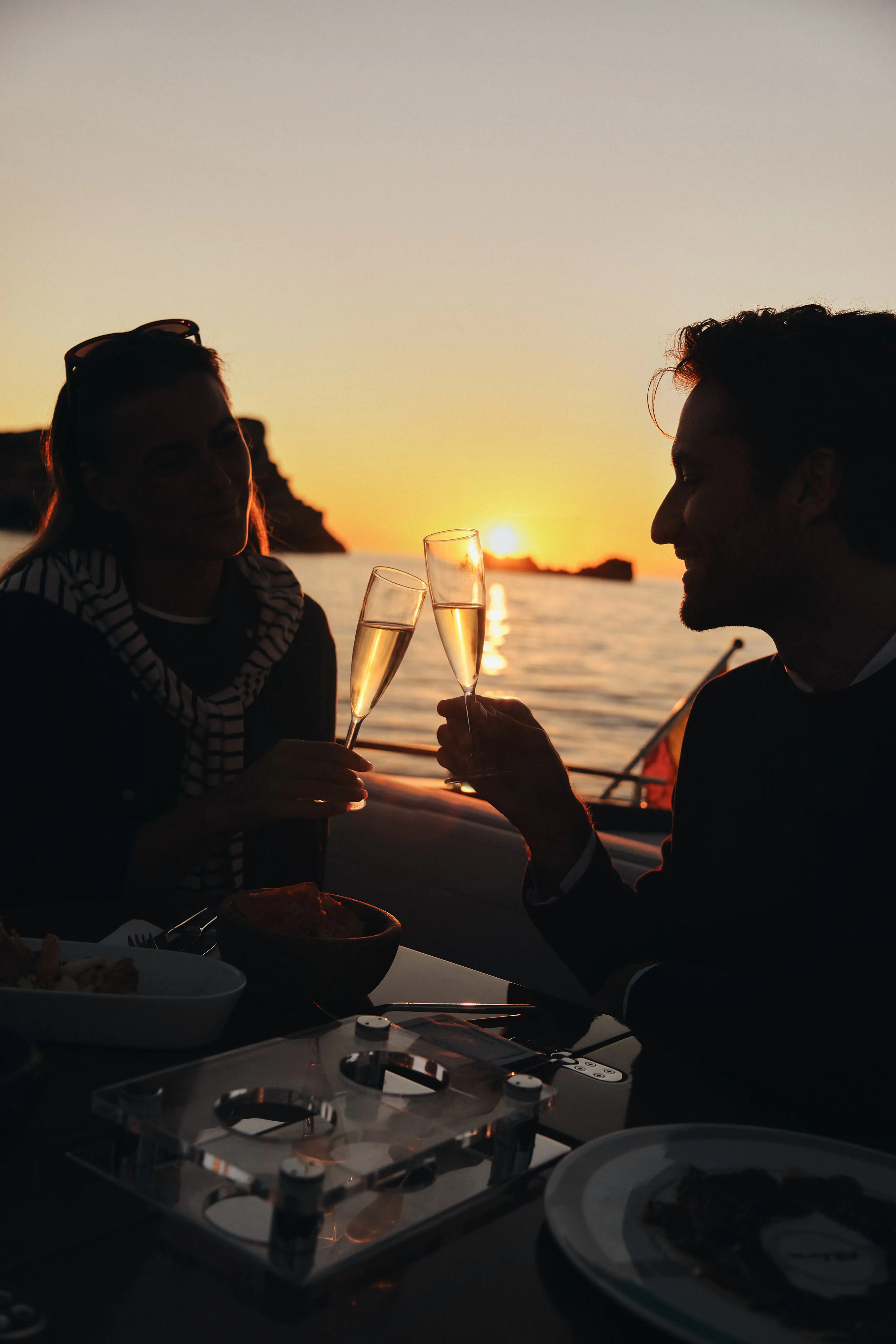 Pareja brindando con copas de champán en un barco al atardecer sobre el mar Mediterráneo.