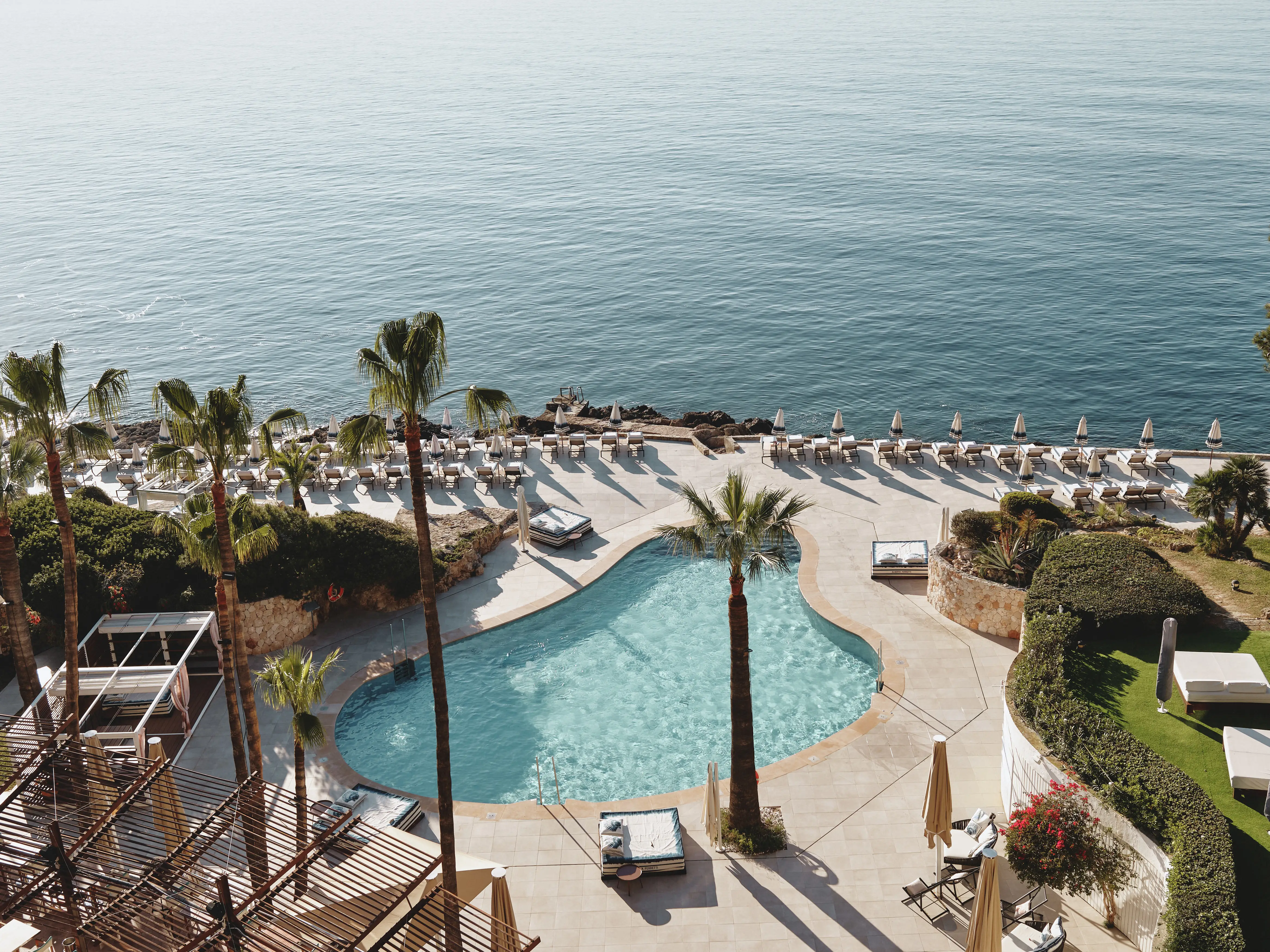 Vista aérea de un resort de lujo junto al mar, con piscina, palmeras y hamacas bajo el sol.