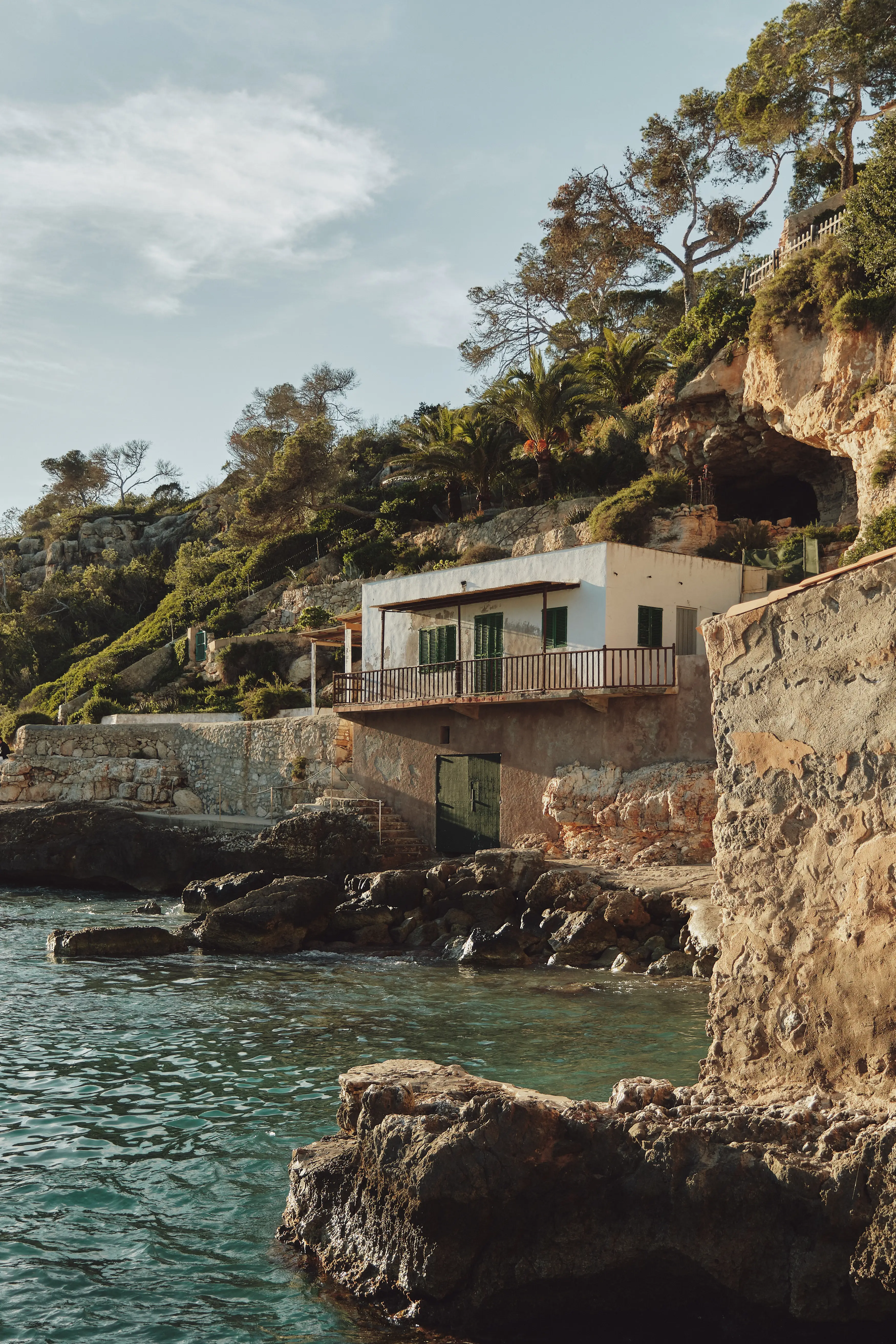 Casita blanca mediterránea con balcón y contraventanas verdes, construida en un acantilado rocoso junto al mar turquesa.
