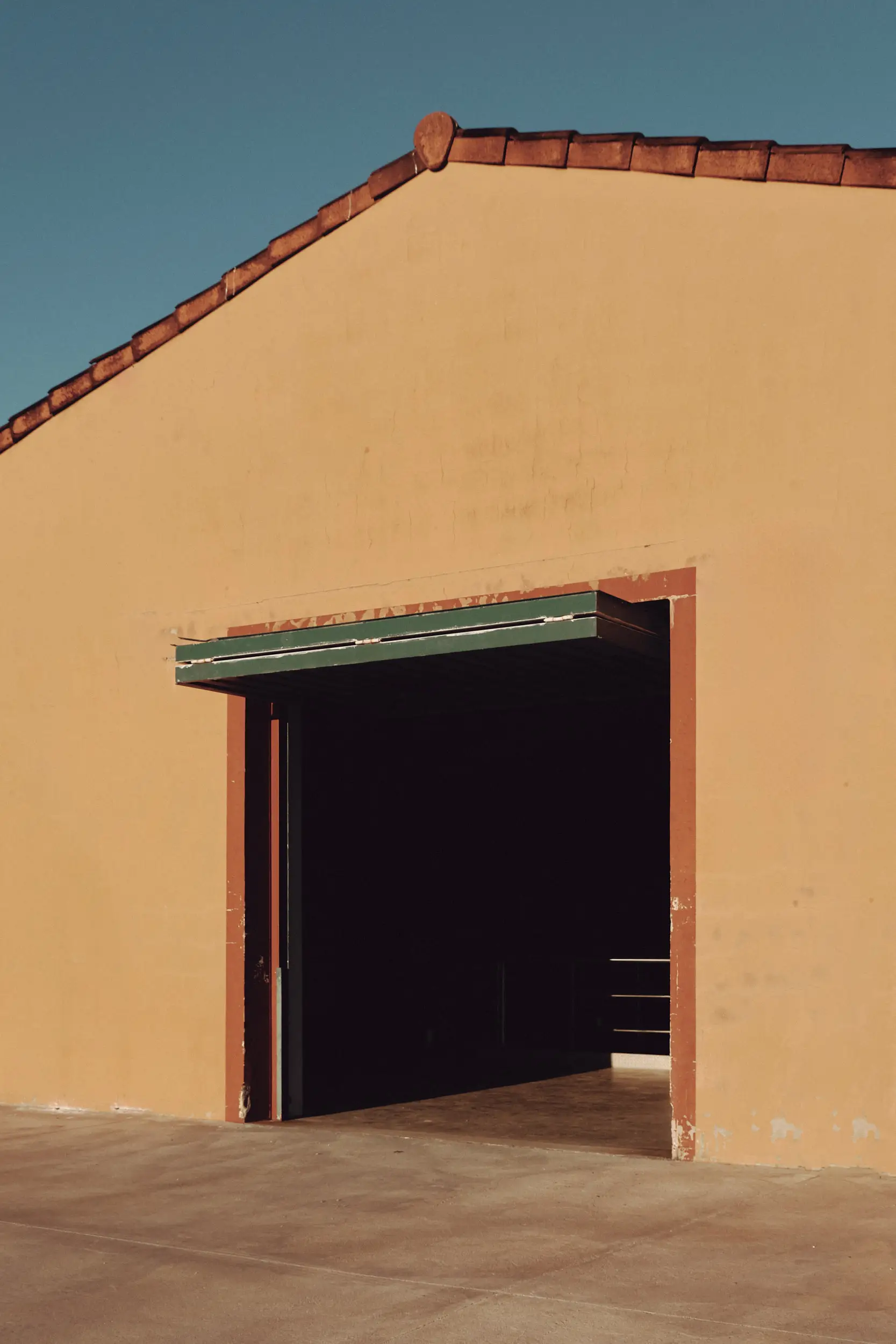 Fachada arquitectónica ocre con tejado de teja y entrada oscura, bañada por el sol en un día claro.