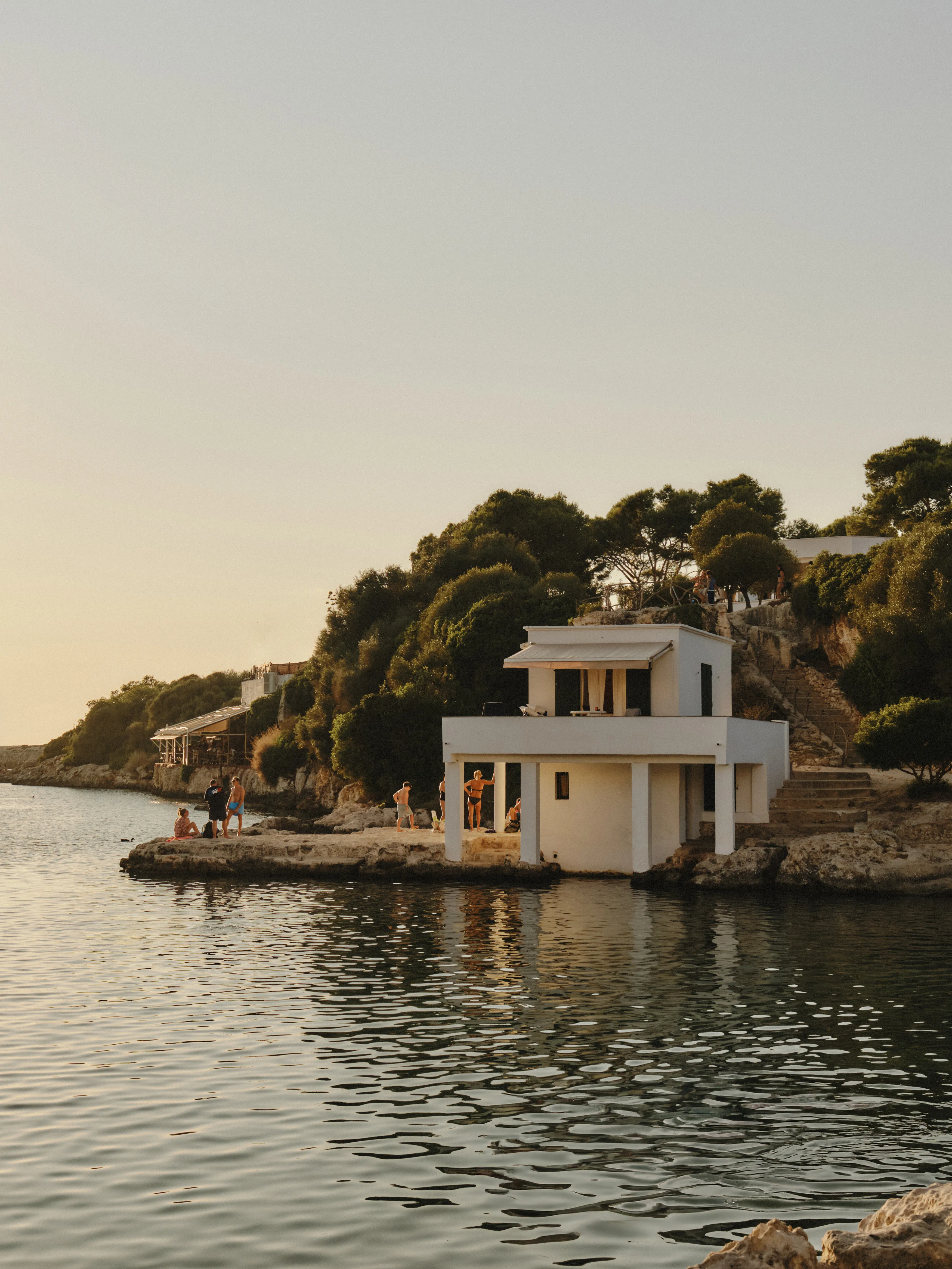 Gente disfrutando de un atardecer mediterráneo junto a una casa blanca en la costa rocosa de una cala.