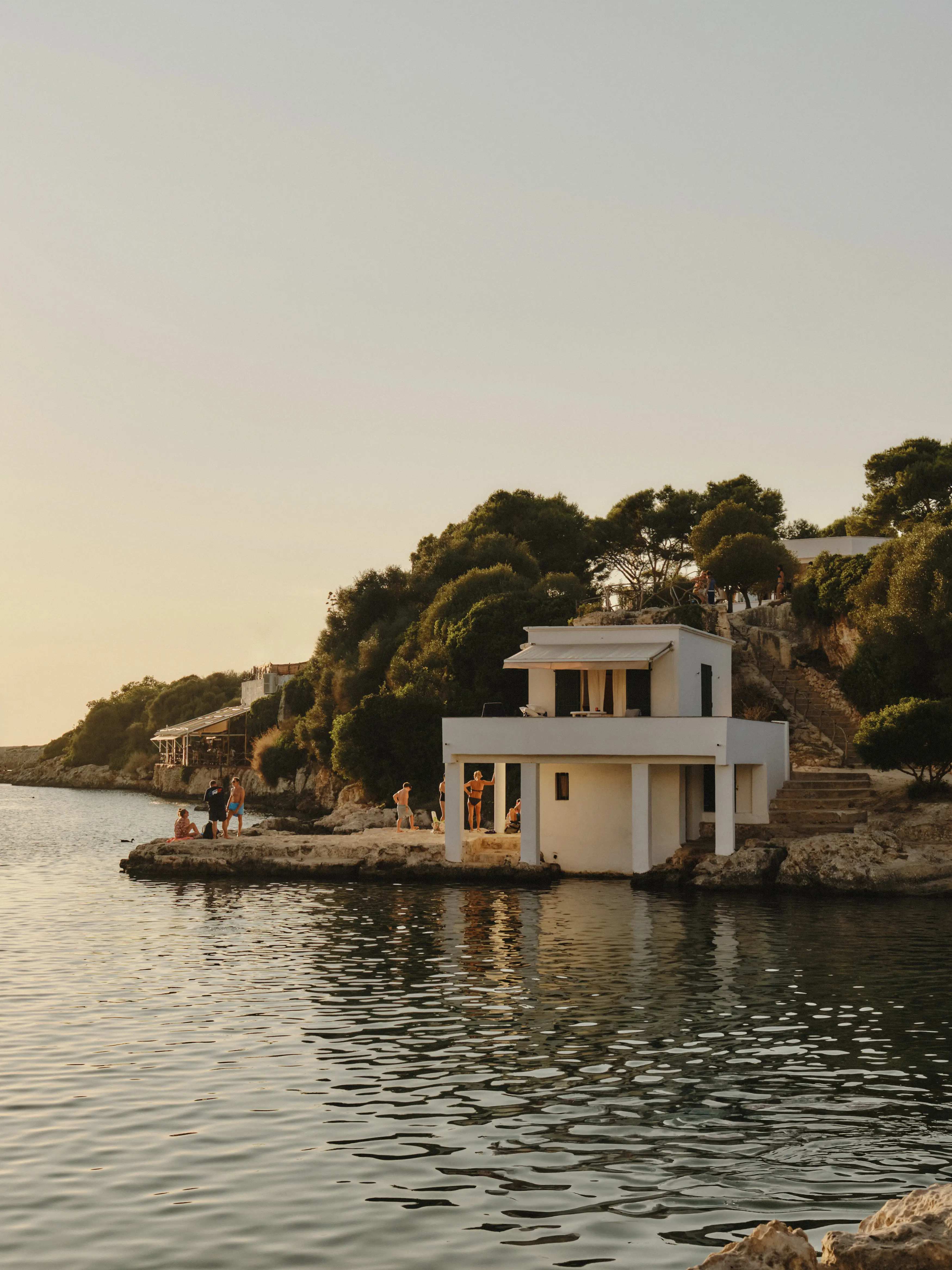Gente disfrutando de un atardecer mediterráneo junto a una casa blanca en la costa rocosa de una cala.