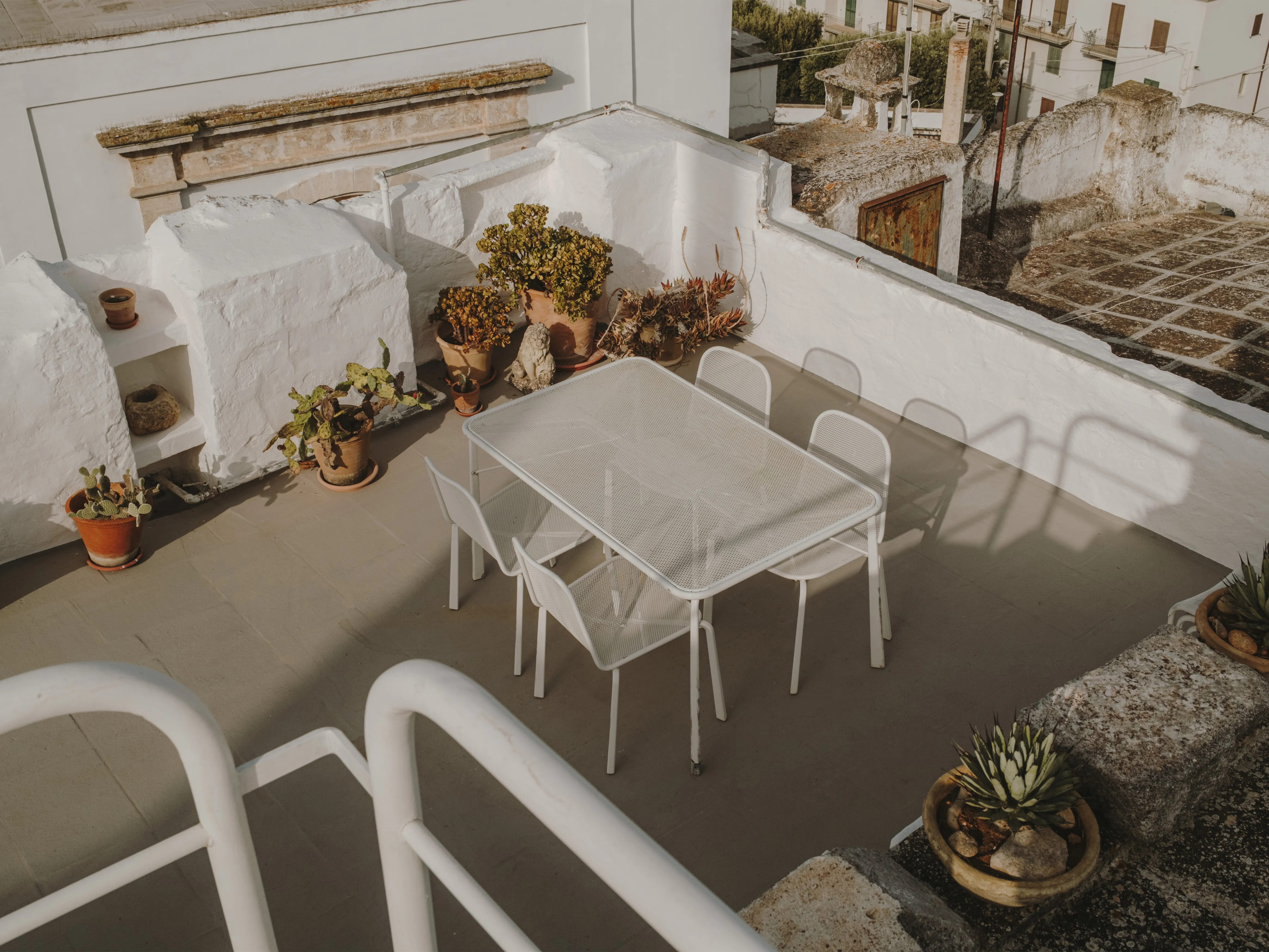 Terraza mediterránea en azotea con mesa y sillas blancas, rodeada de plantas en macetas y vistas a un pueblo.