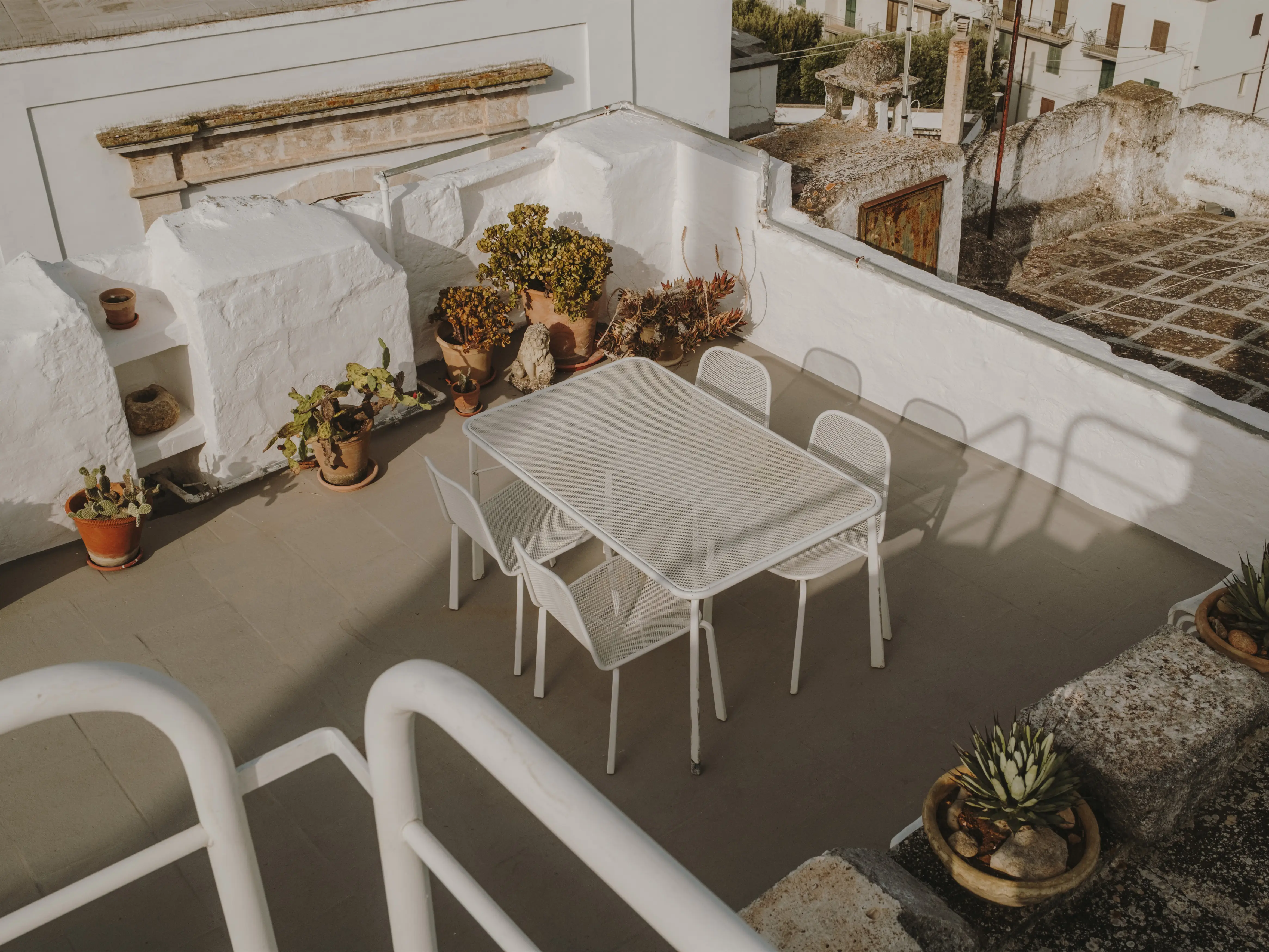 Terraza mediterránea en azotea con mesa y sillas blancas, rodeada de plantas en macetas y vistas a un pueblo.