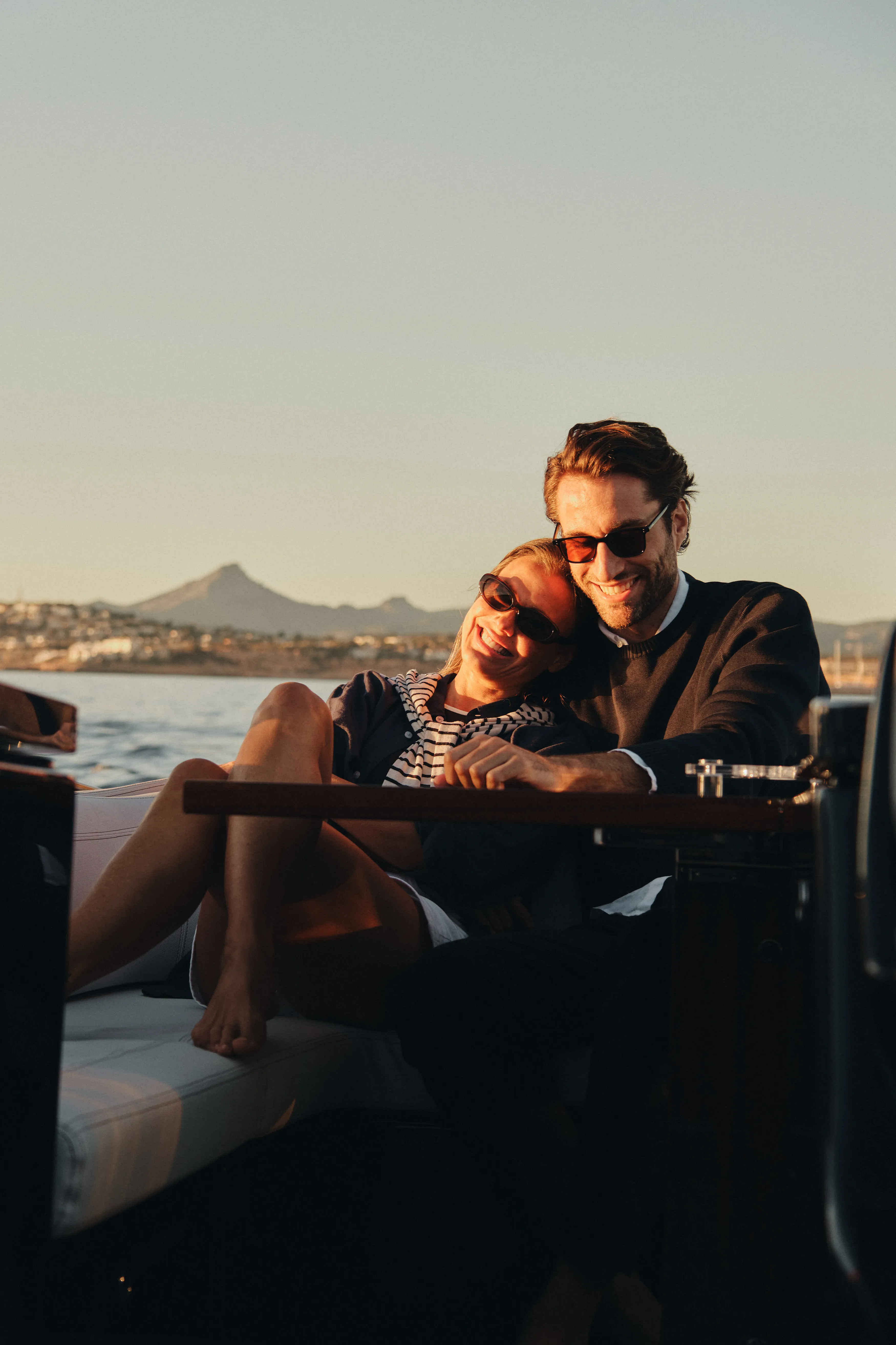 Pareja sonriente disfrutando del atardecer en un barco, con un paisaje costero montañoso de fondo.