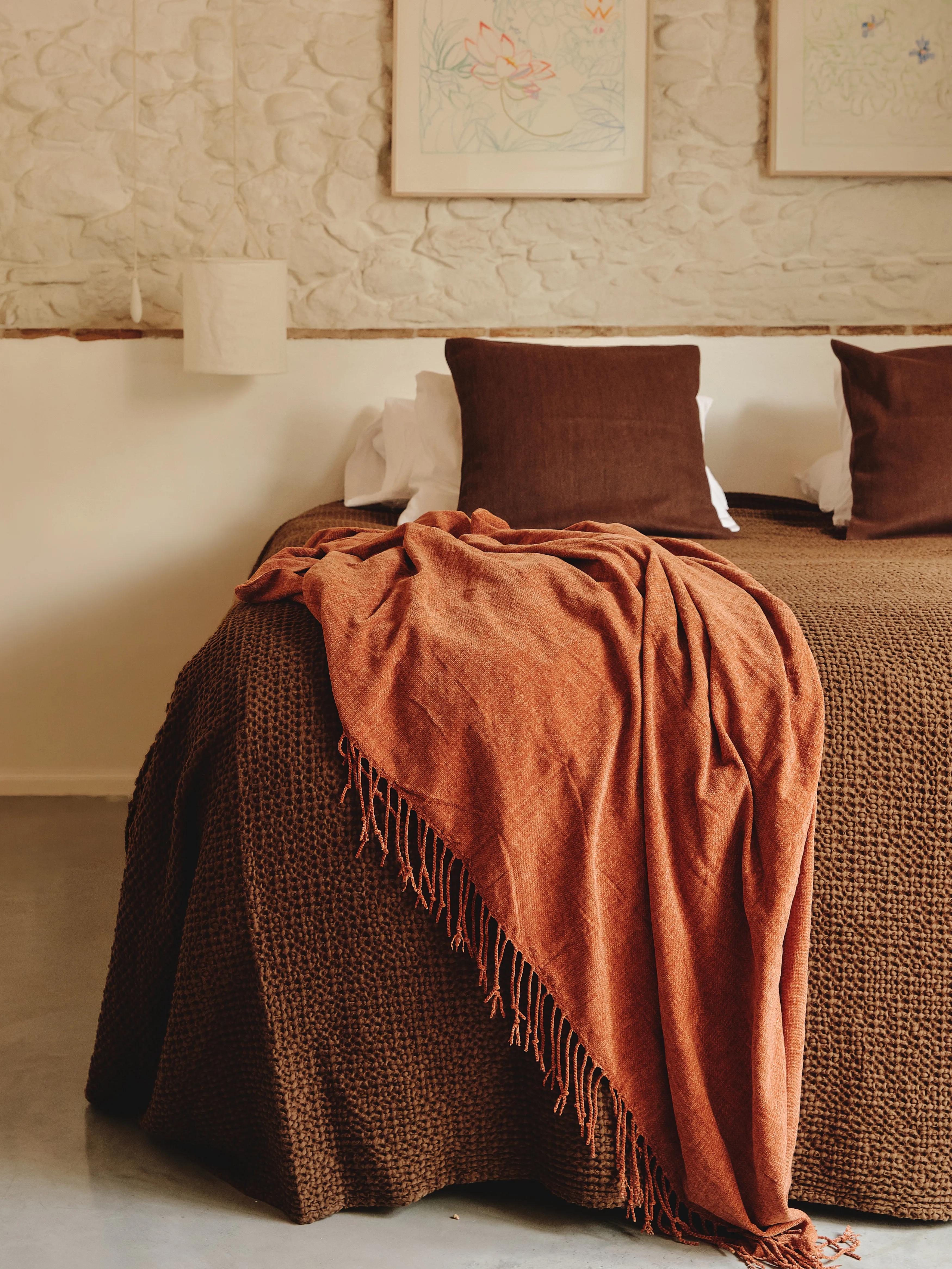 Dormitorio acogedor con pared de piedra, arte botánico y ropa de cama cálida en tonos tierra y rojizos.
