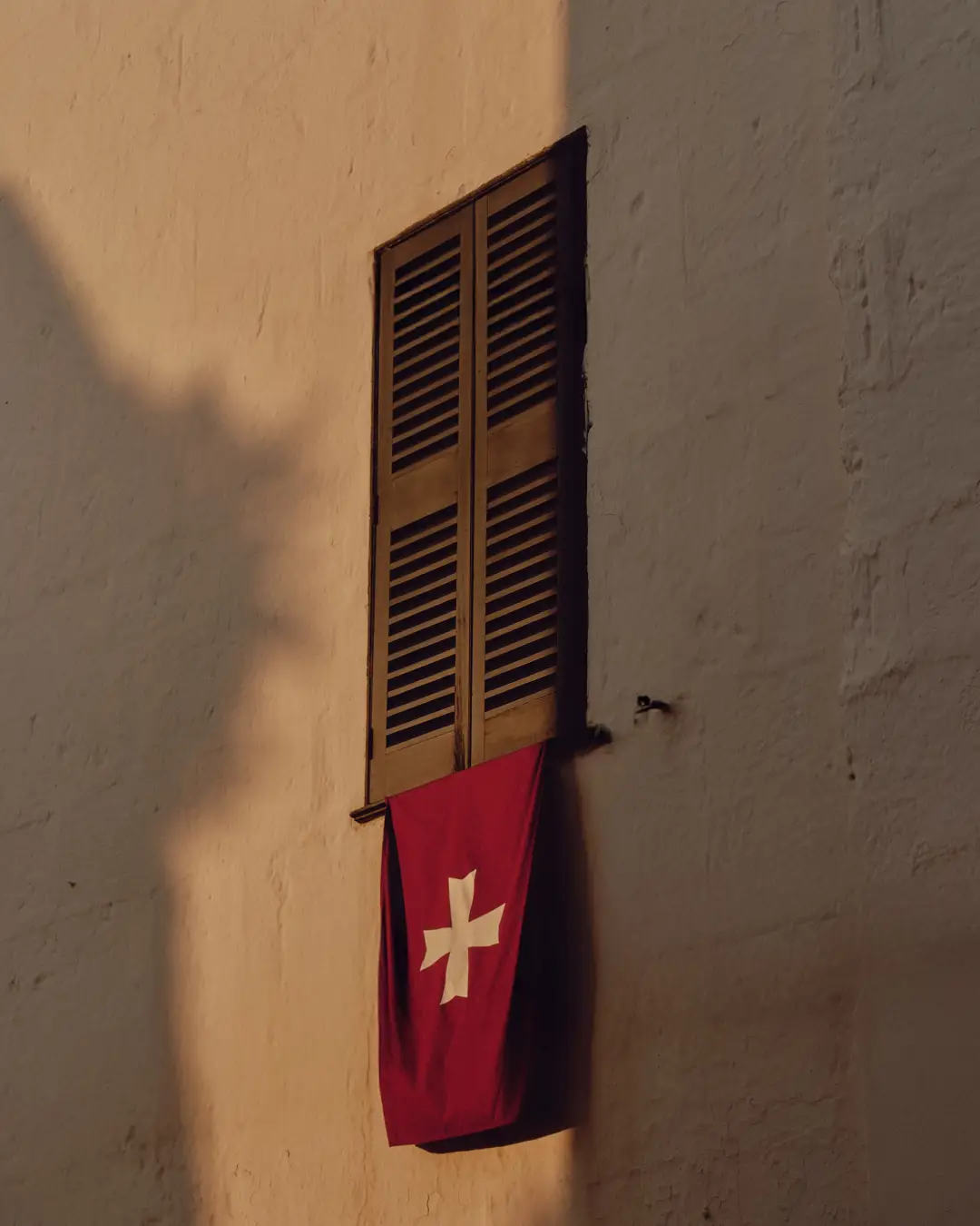 Bandera suiza colgando de una ventana con persianas cerradas en una fachada con luz cálida y sombras.