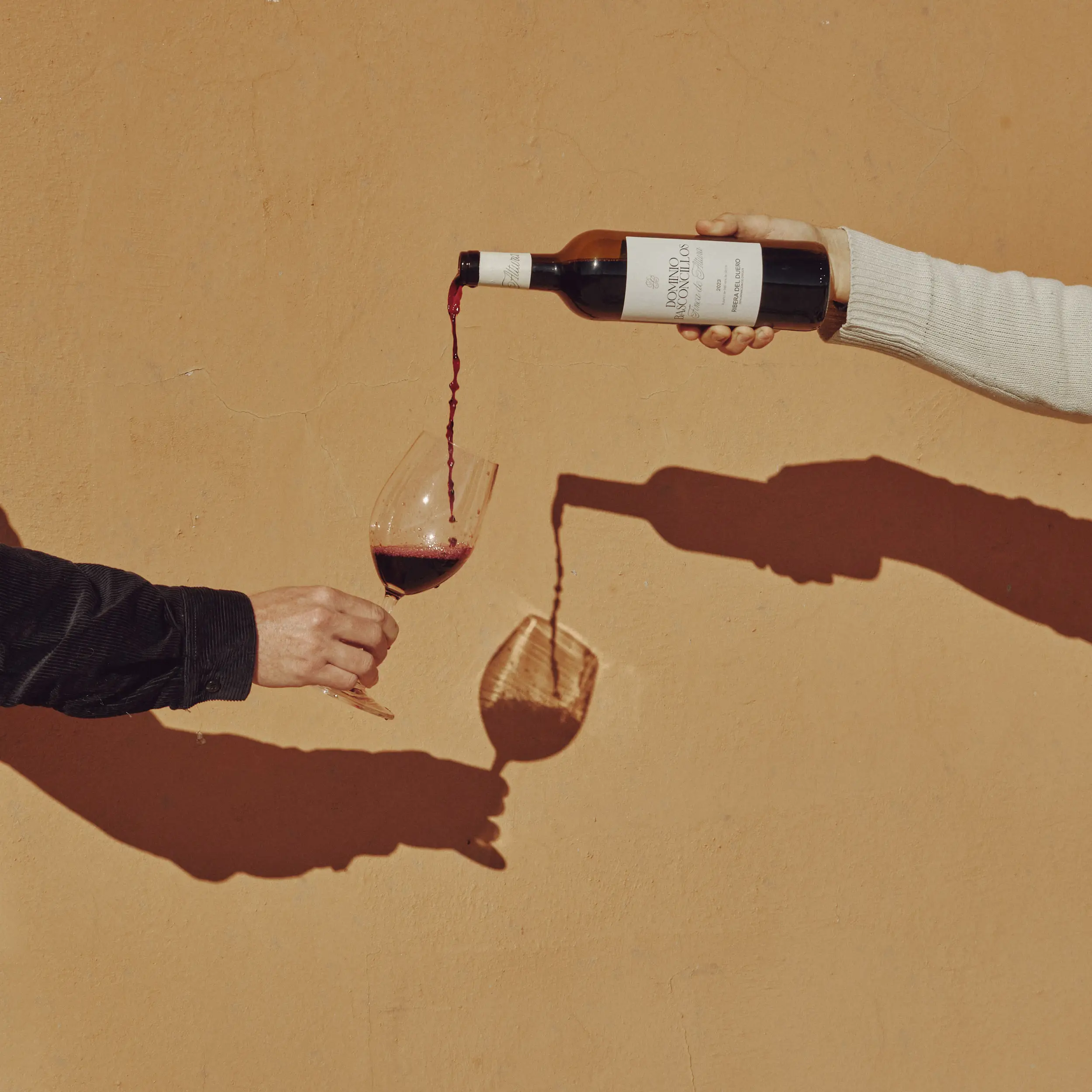 Dos manos vierten vino tinto de una botella a una copa, con sombras proyectadas en una pared ocre.