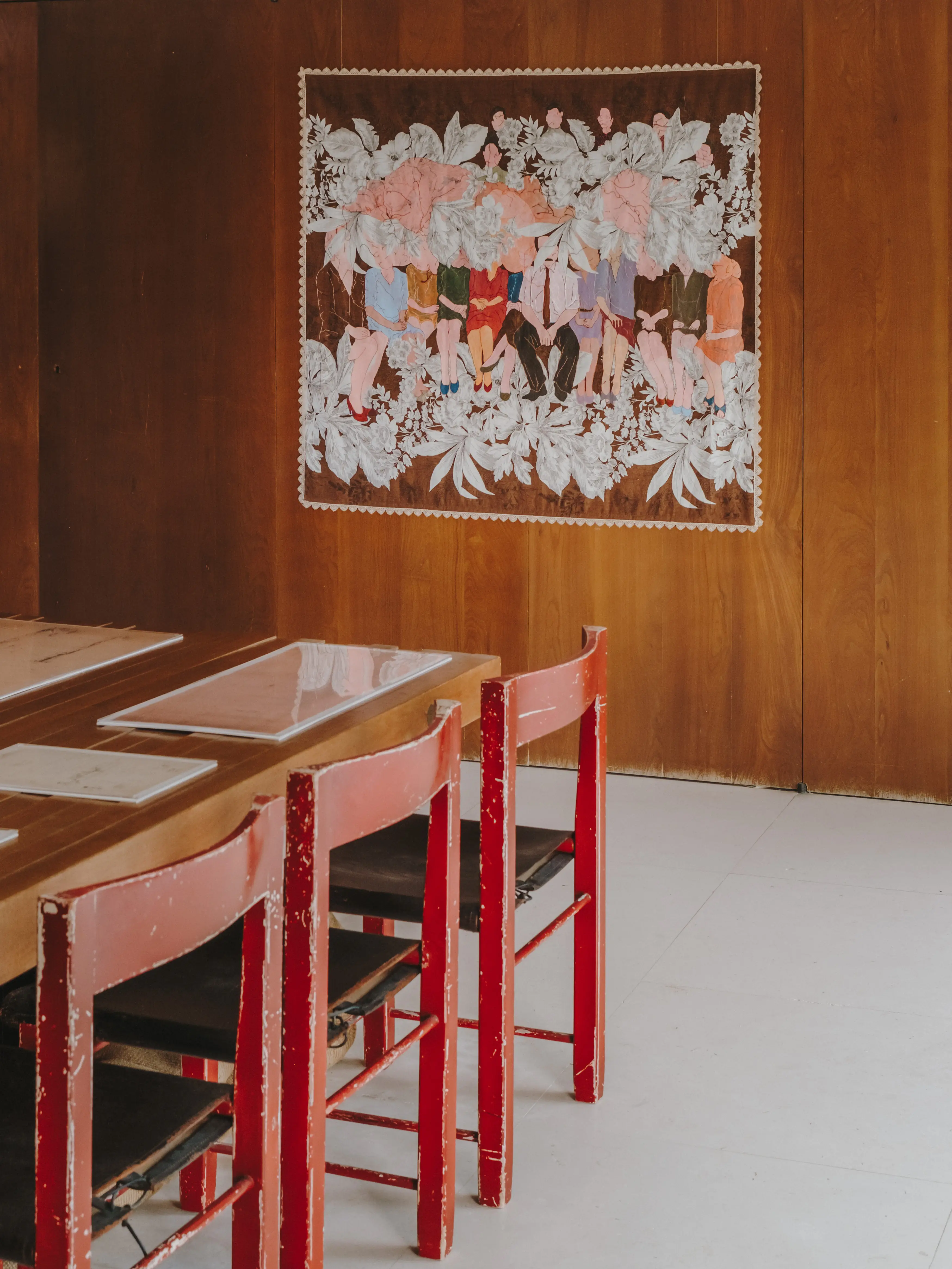 Interior artístico con una gran obra textil en pared de madera, sillas rojas desgastadas y mesa de madera.