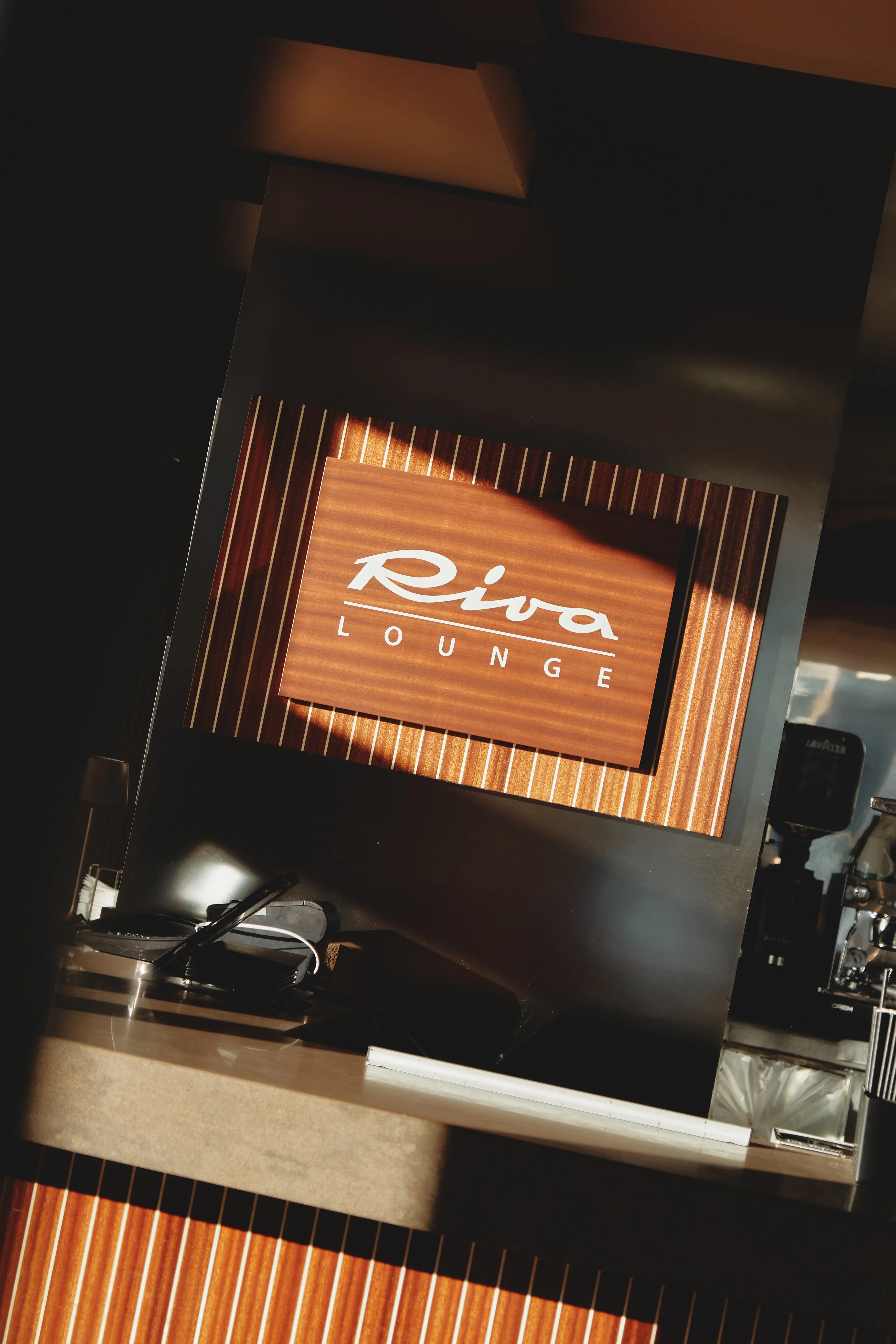 Rótulo de madera "Riva Lounge" iluminado por el sol, sobre una barra moderna en un interior acogedor.