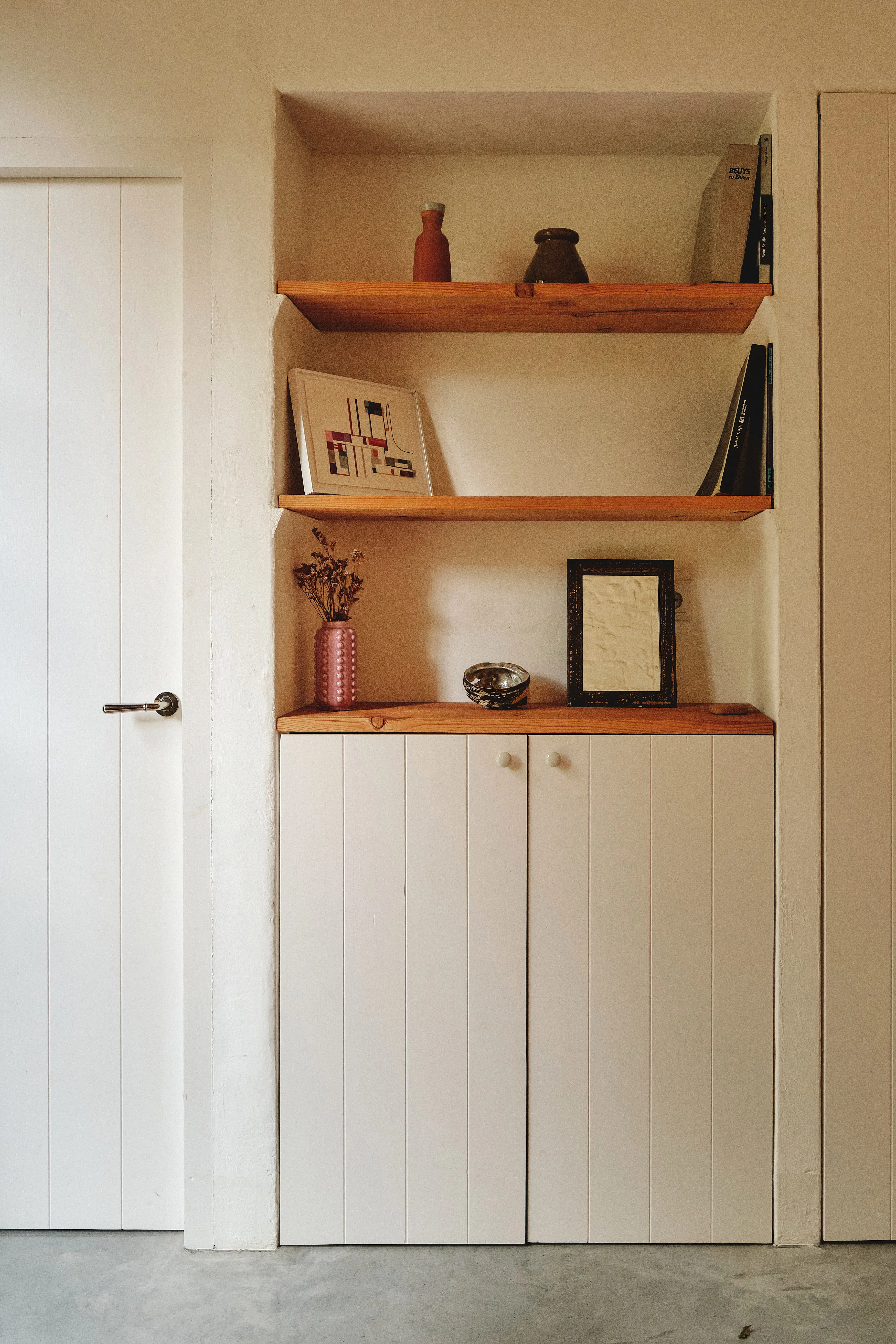Nicho empotrado con estantes de madera y armarios bajos blancos, decorado con arte, libros y objetos artesanales.