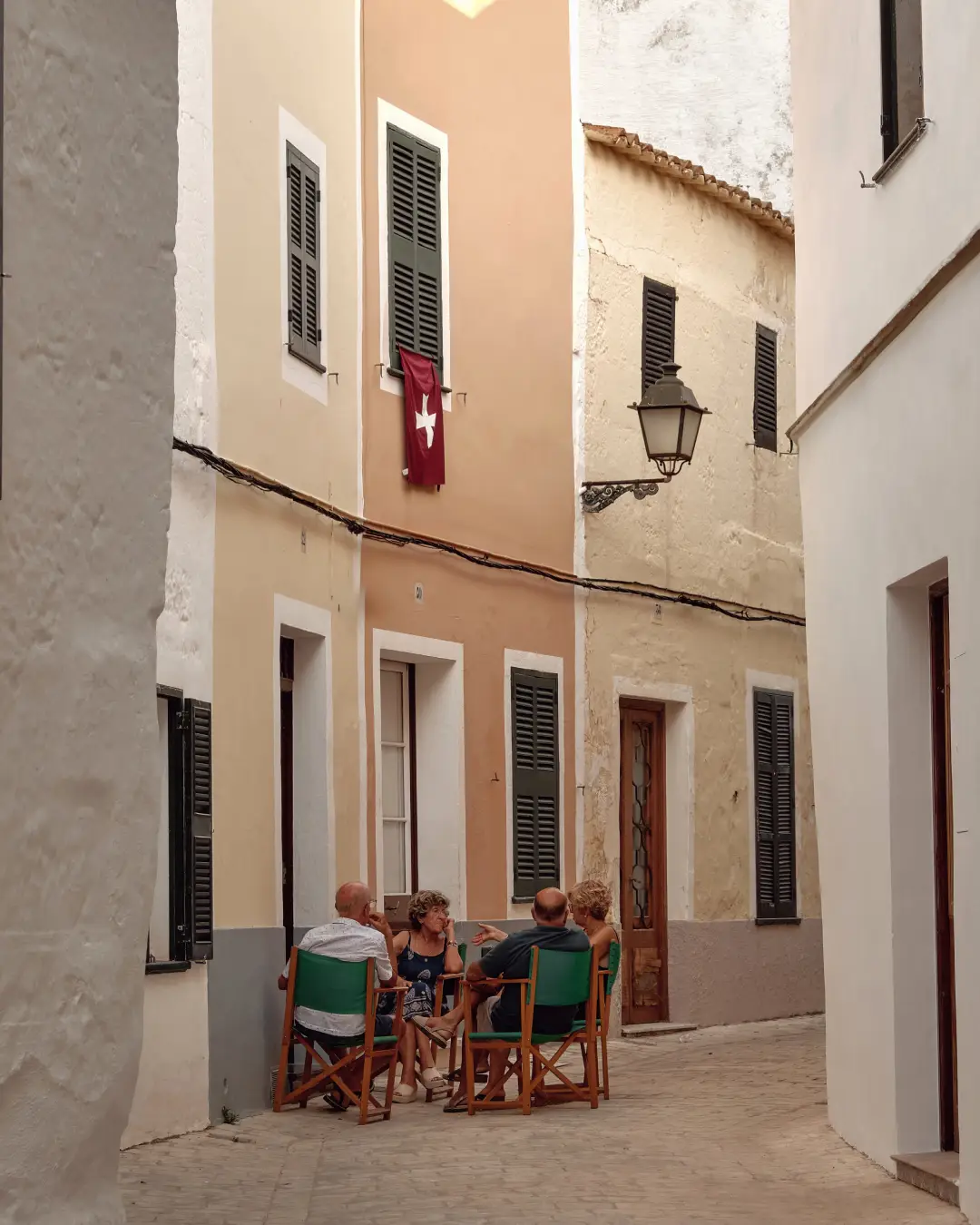 Cuatro personas conversan relajadamente en una estrecha calle mediterránea rodeada de edificios con persianas tradicionales.