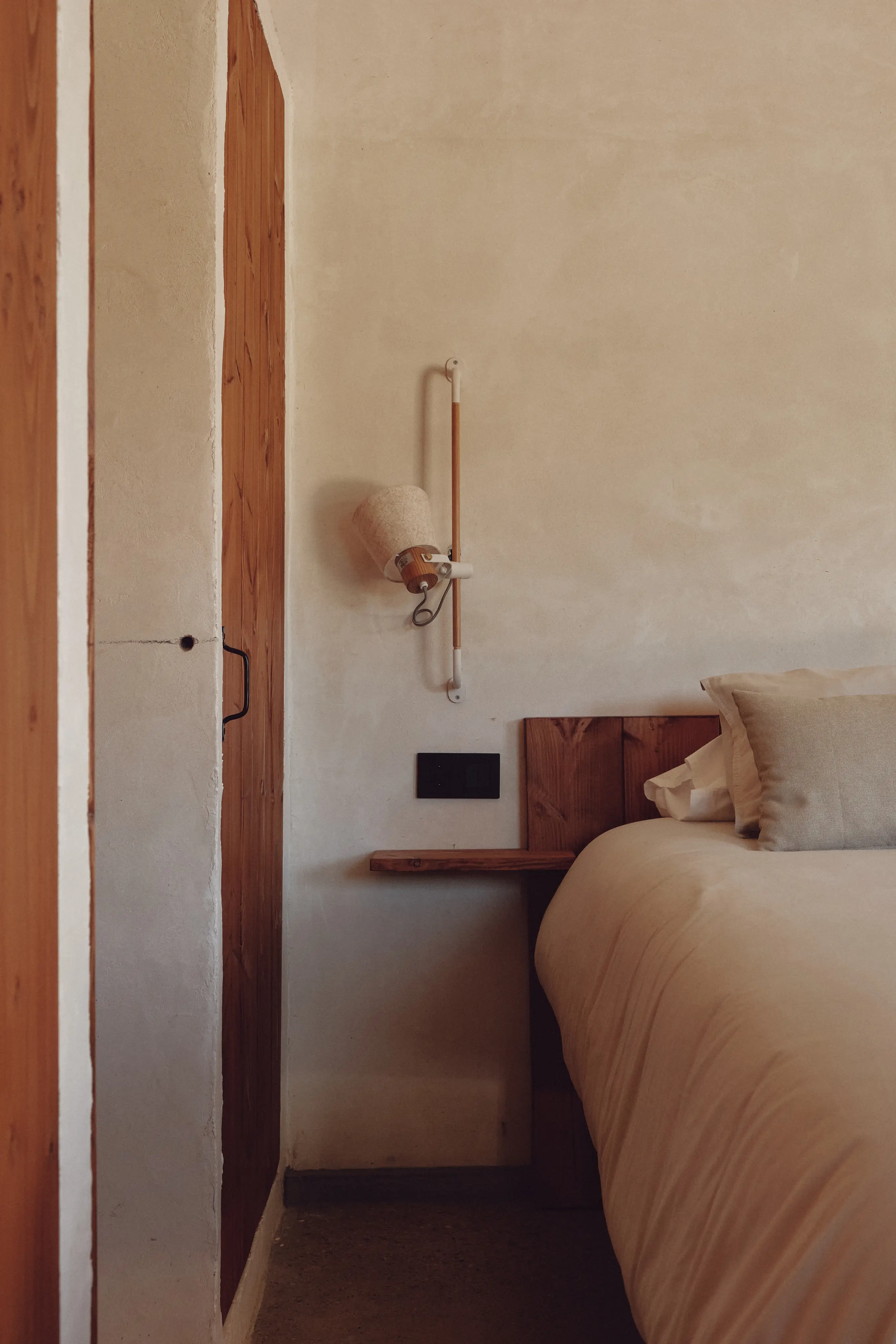 Acogedor detalle de dormitorio mediterráneo con cama, cabecero y mesilla de madera, y una lámpara de pared.