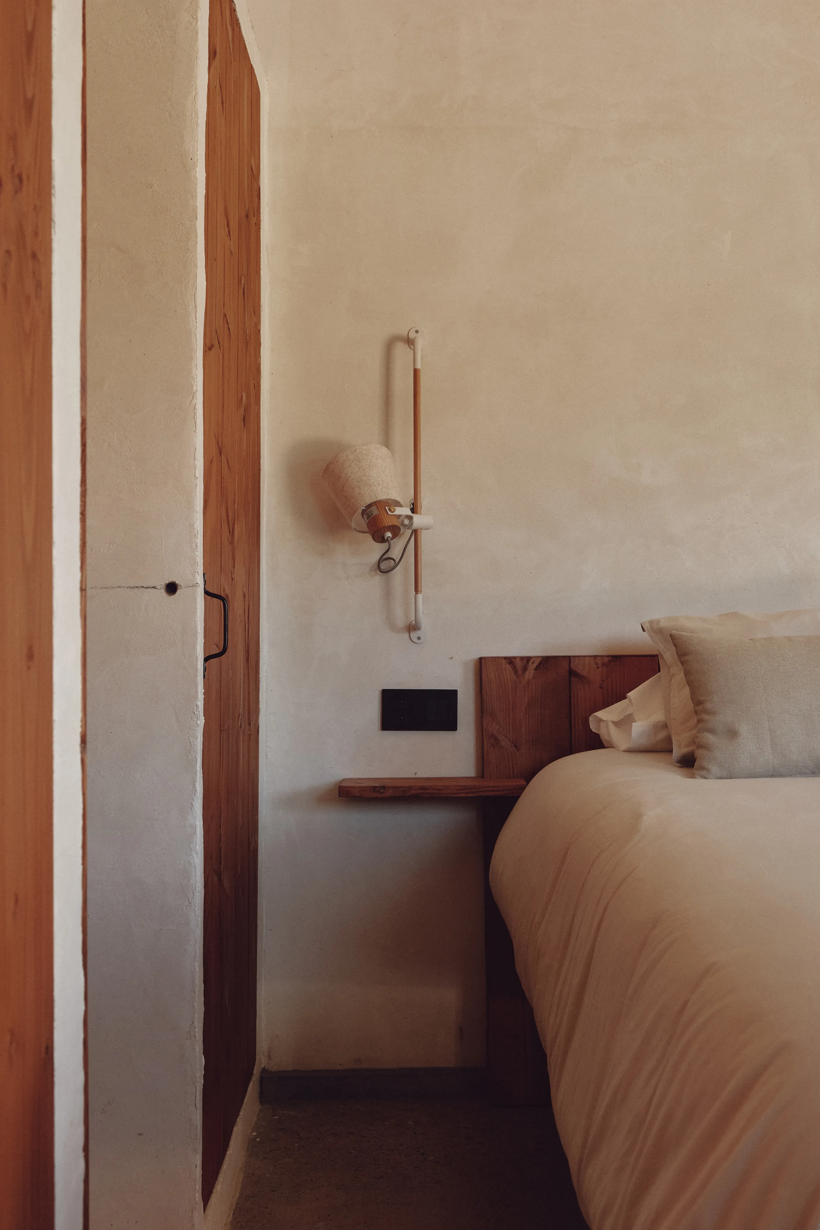 Acogedor detalle de dormitorio mediterráneo con cama, cabecero y mesilla de madera, y una lámpara de pared.