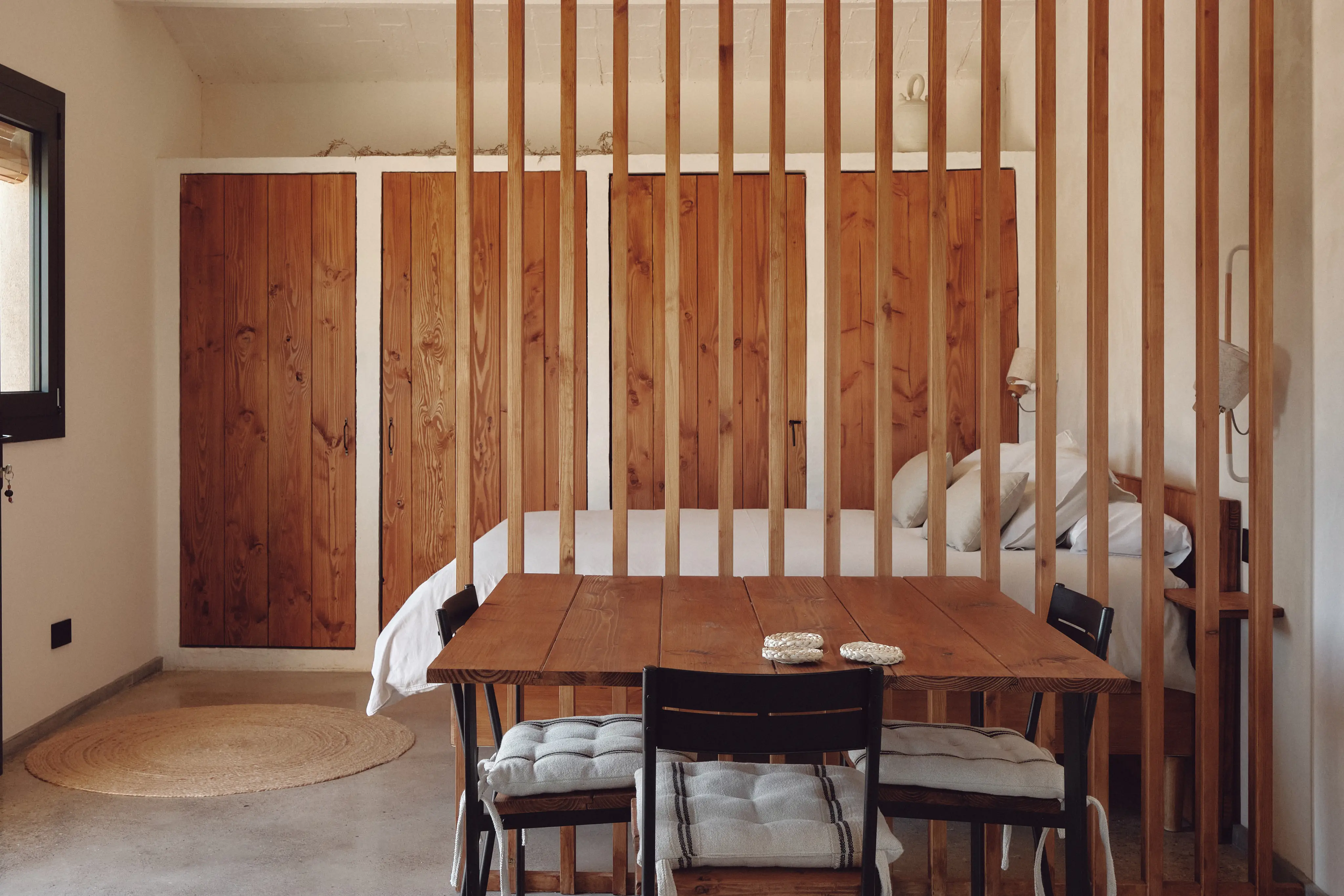 Interior cálido con mesa de madera, sillas y cama, separados por un divisor de listones. Estilo rústico y funcional.