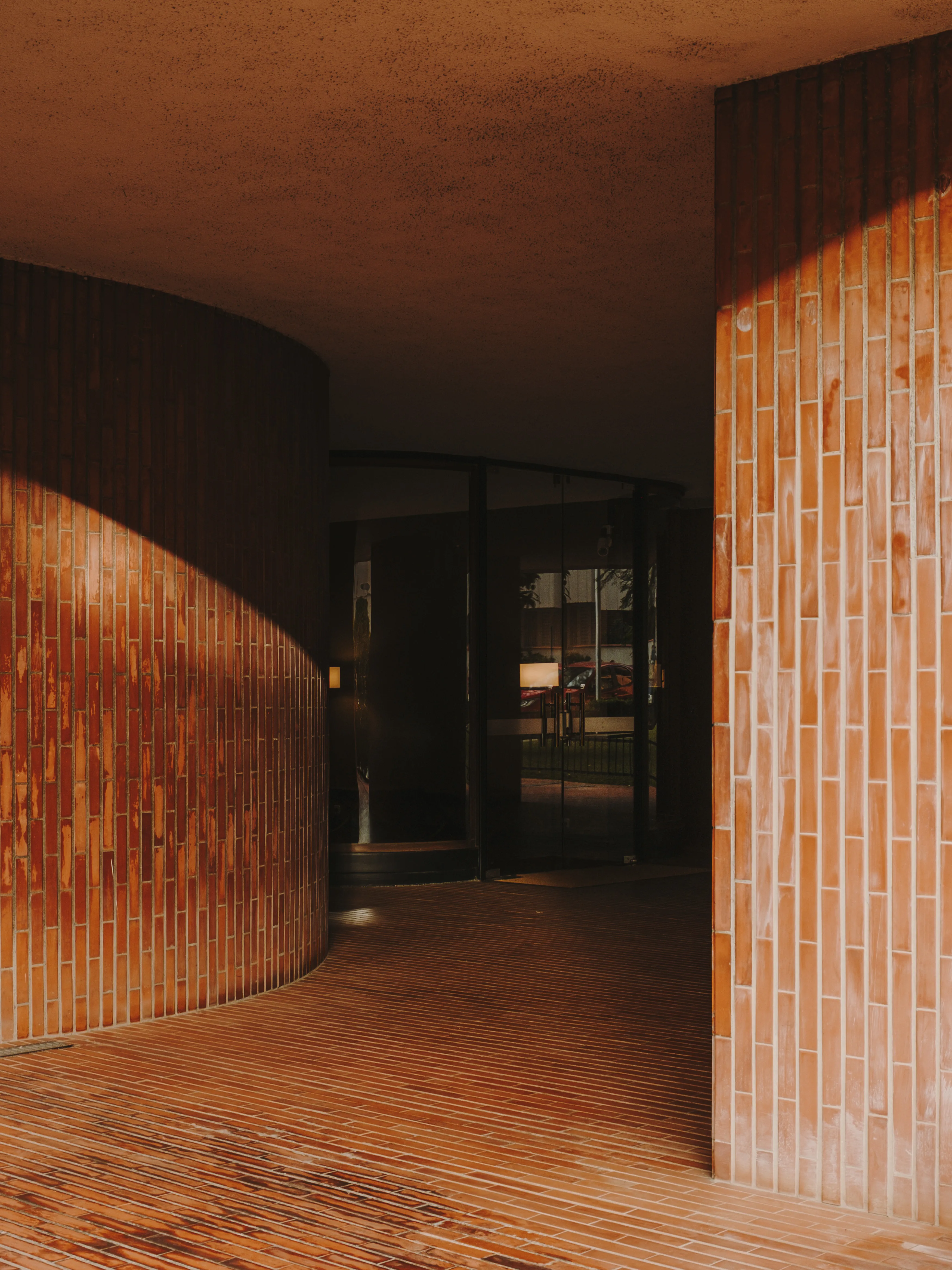 Pasillo arquitectónico con paredes y suelo de ladrillo curvado y recto, iluminado por el sol, con entrada de cristal oscuro al fondo.