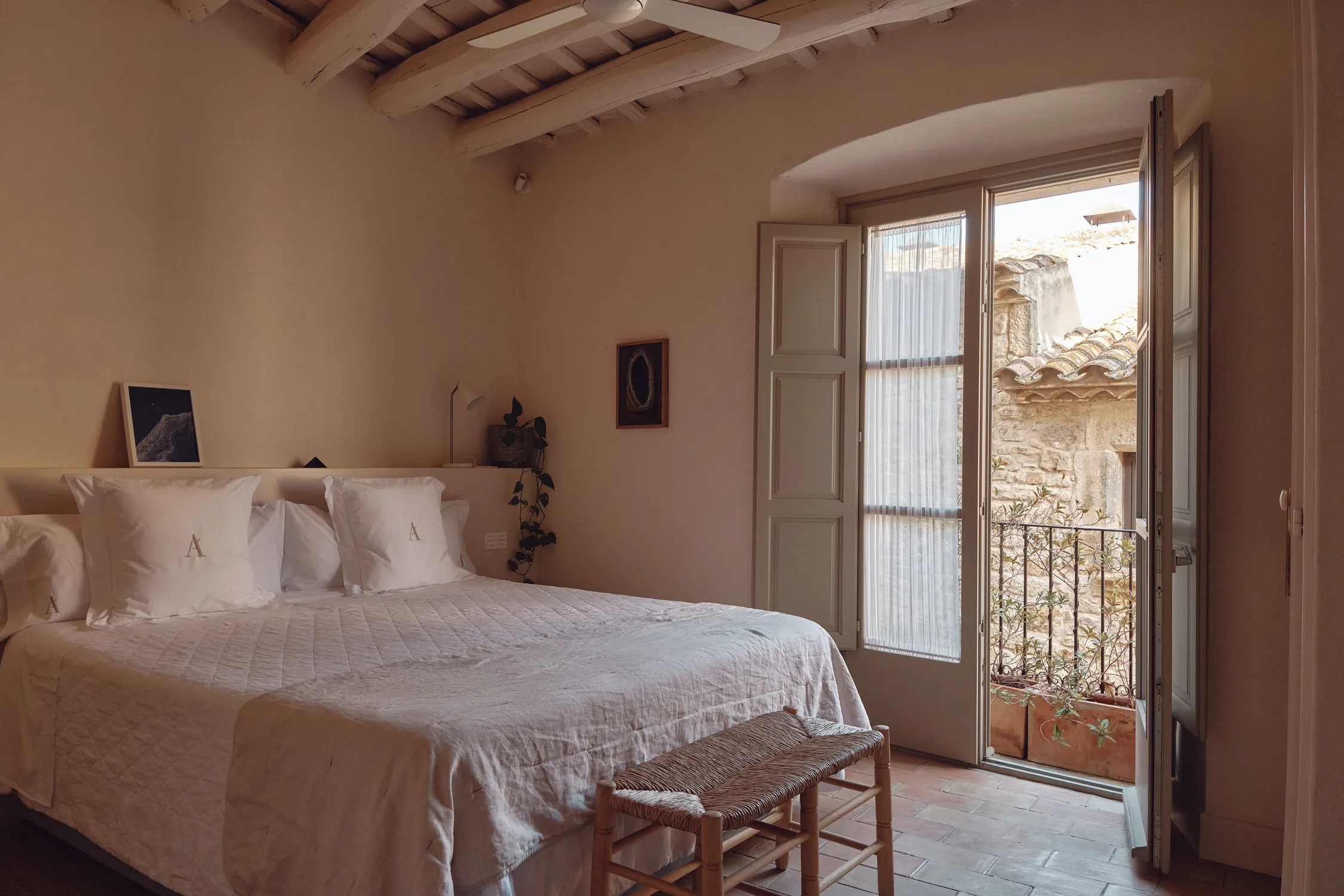 Acogedor dormitorio mediterráneo con cama doble, techo de vigas de madera, y balcón abierto a una fachada de piedra tradicional.